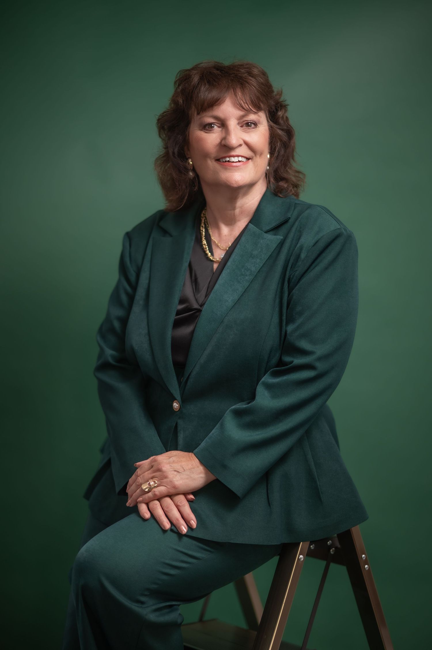 a woman with dark curly hair in an emerald green suit sits in front of an emerald green studio background and smiles for a portrait