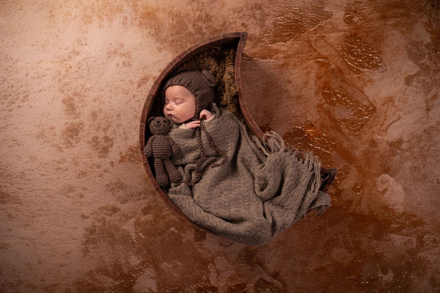 Newborn baby sleeping peacefully in a crescent moon prop, wrapped in gray knit blanket with tiny teddy bear.