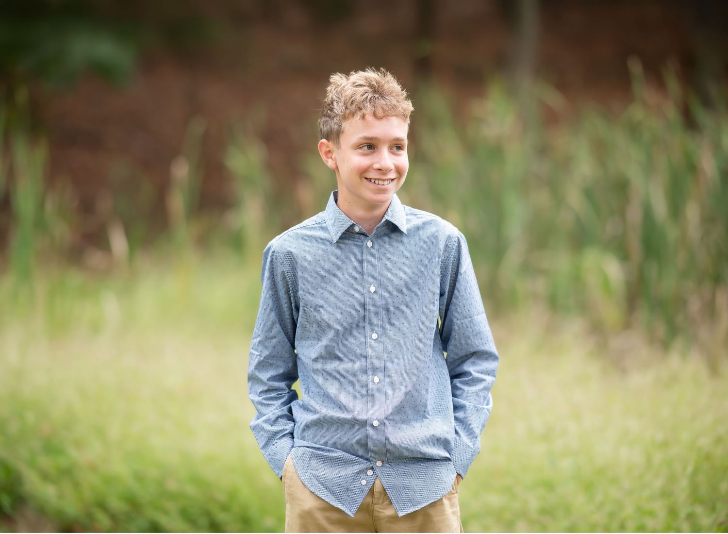 a young man in a blue dress shirt stands in front of tall grass at the edge of a pond and laughs; shot in Morgantown, WV