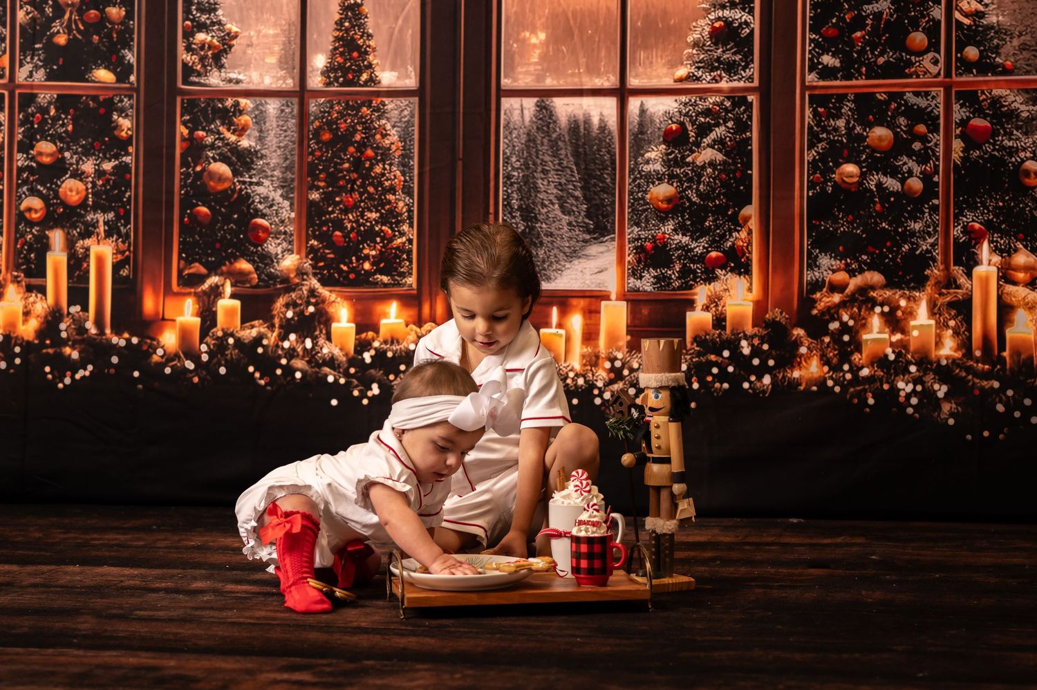 two little siblings in Christmas pajamas sit on the wood floor with a tray of hot cocoa and cookies, and Christmas trees in the background