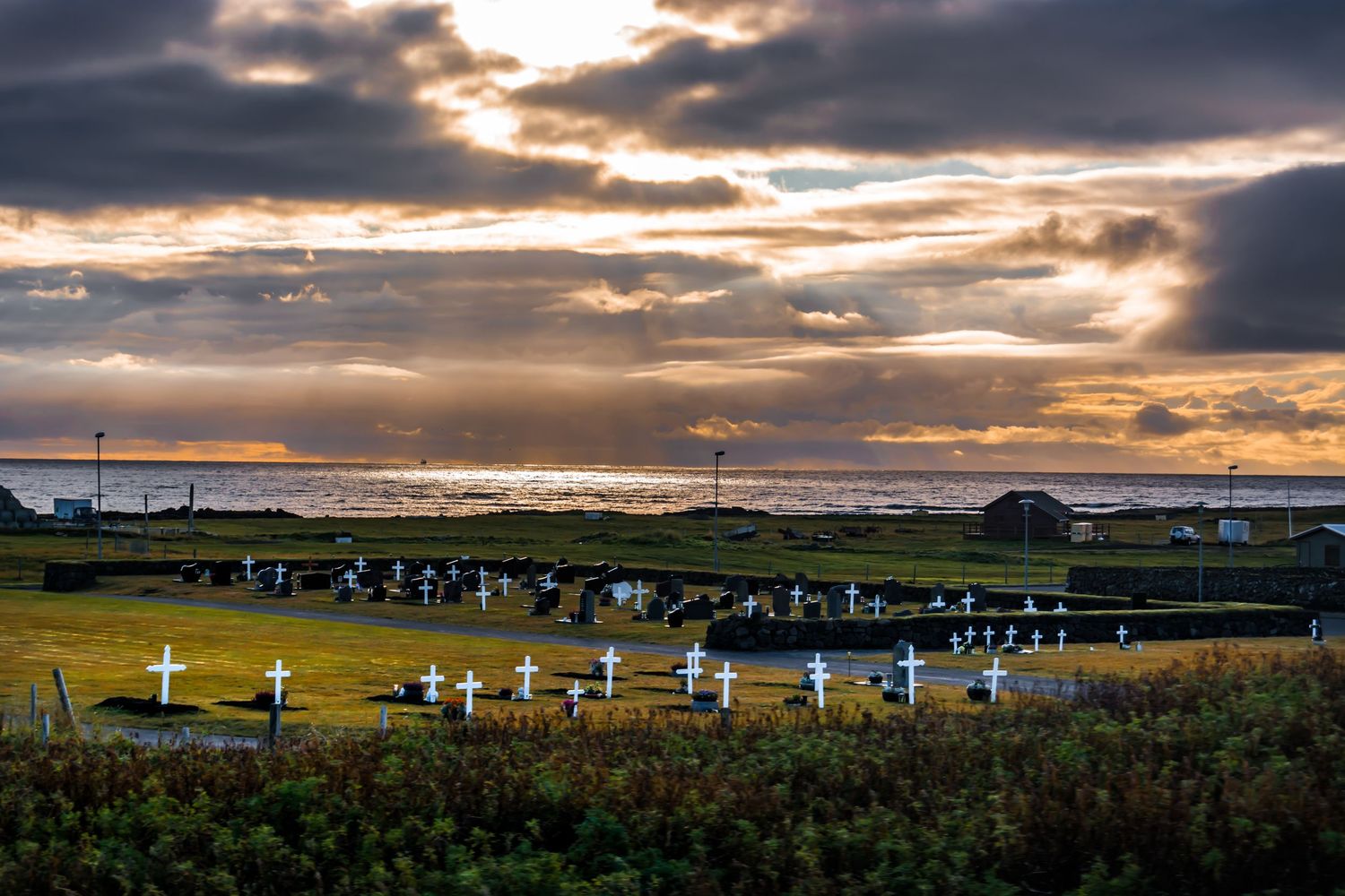 Sunset over a coastal cemetery with white crosses on green grass, dramatic cloudy sky with rays of light over the ocean.