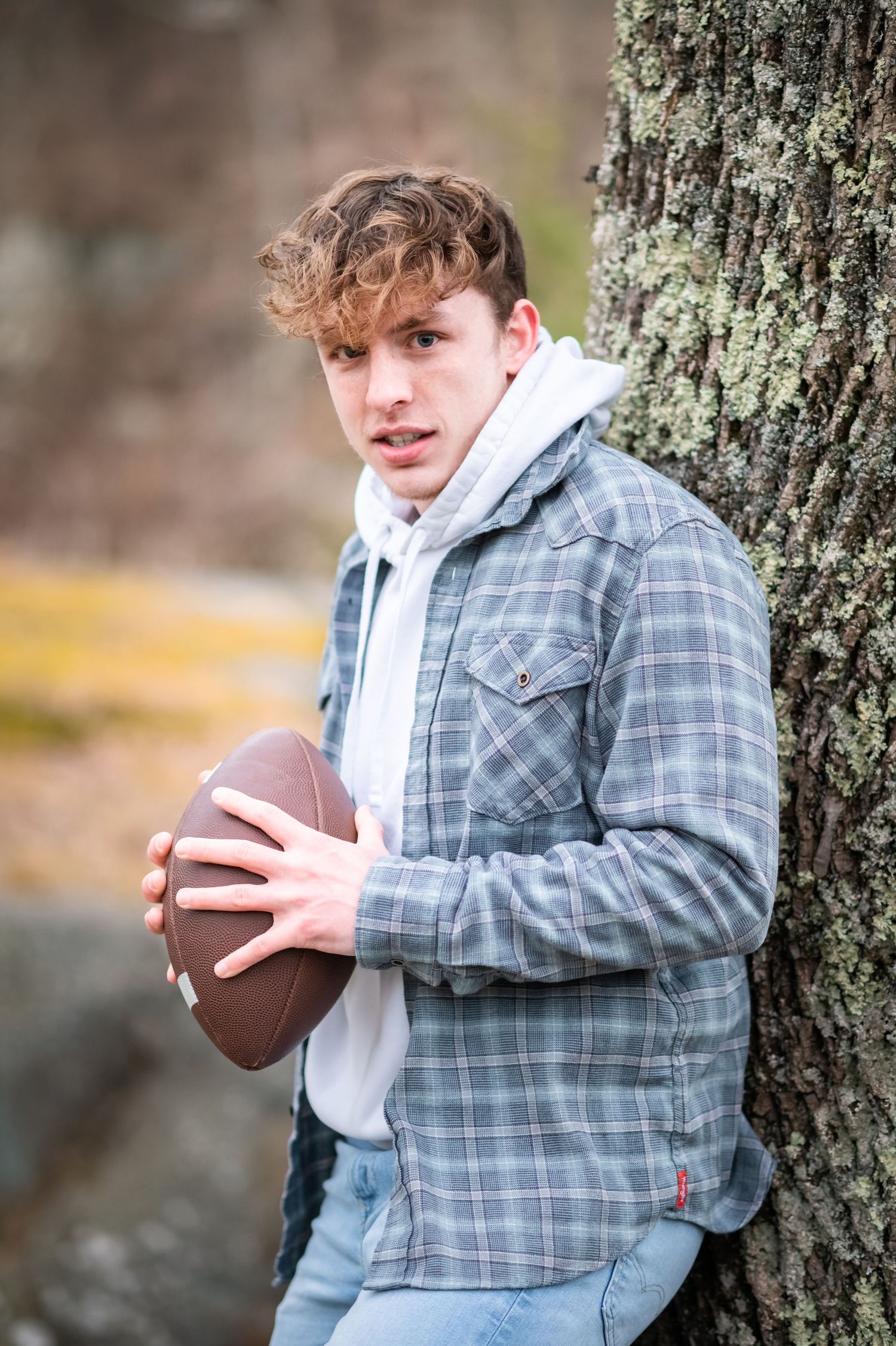 a high school football player in a hoodie and jeans leans against a tree and poses with a football for a senior portrait in Fairmont, WV