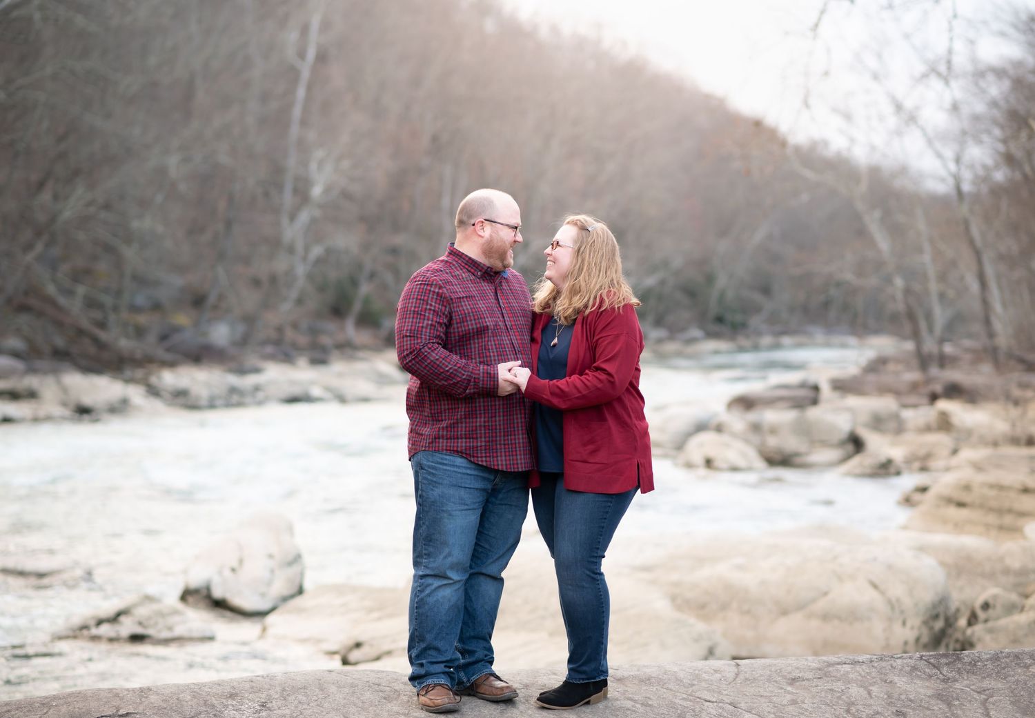 a husband and wife in red sweaters stand together on the riverbank at Valley Falls, WV