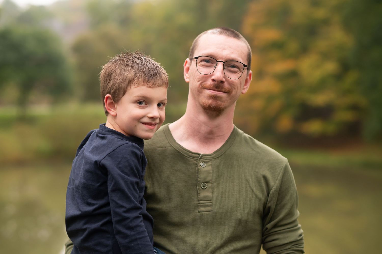 a father holds his young son and they smile for a portrait, with autumn scenery in the background