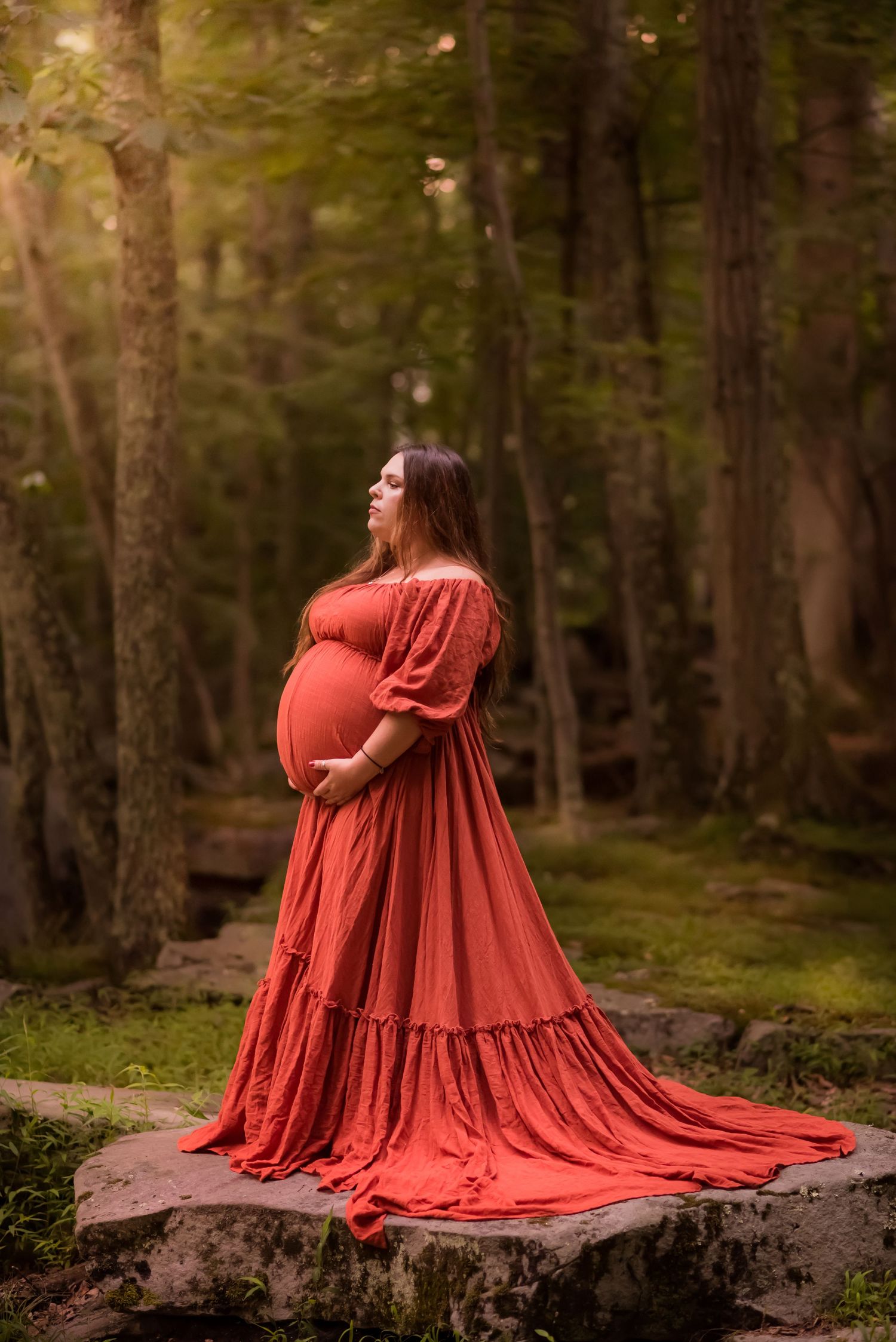 a pregnant woman in a long orange dress with a train stands in a clearing in the woods at Valley Falls State Park in Fairmont, WV
