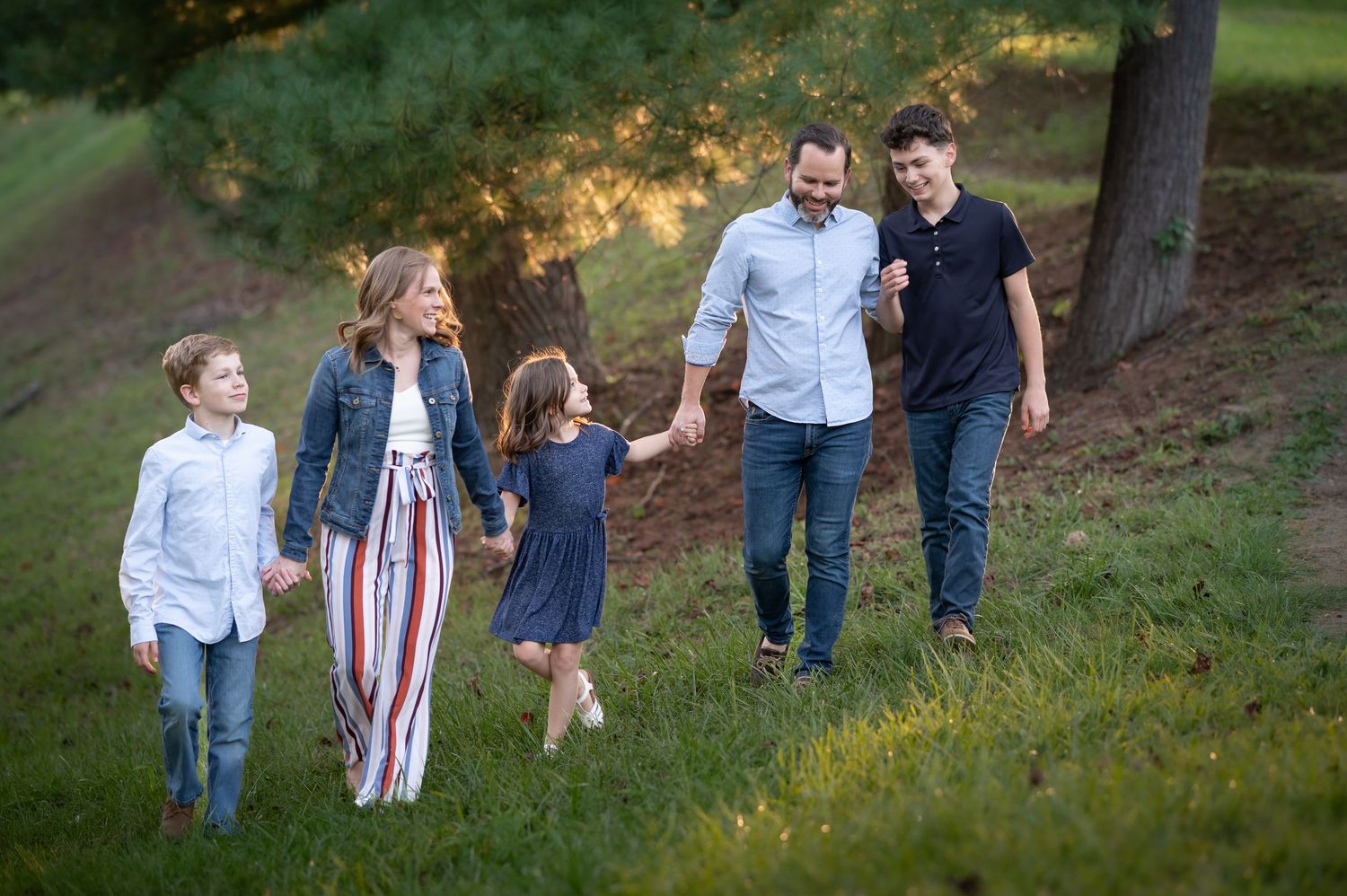 a family of five holds hands and walks laughing down a grassy hillside with sunlit trees in the background; Morgantown, WV