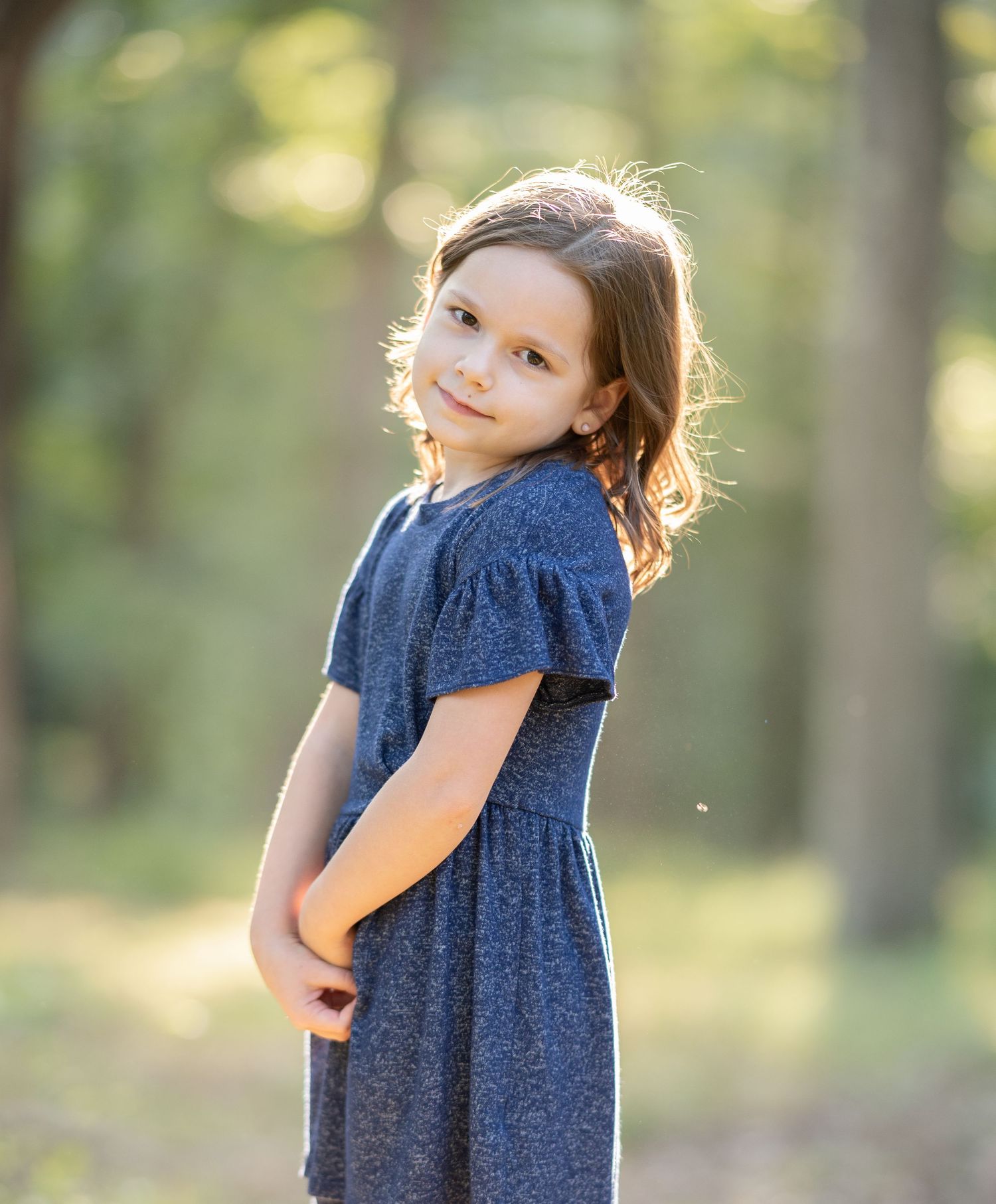 a little girl in a blue dress poses for a portrait with her hands clasped together, and soft green trees blurred in the background