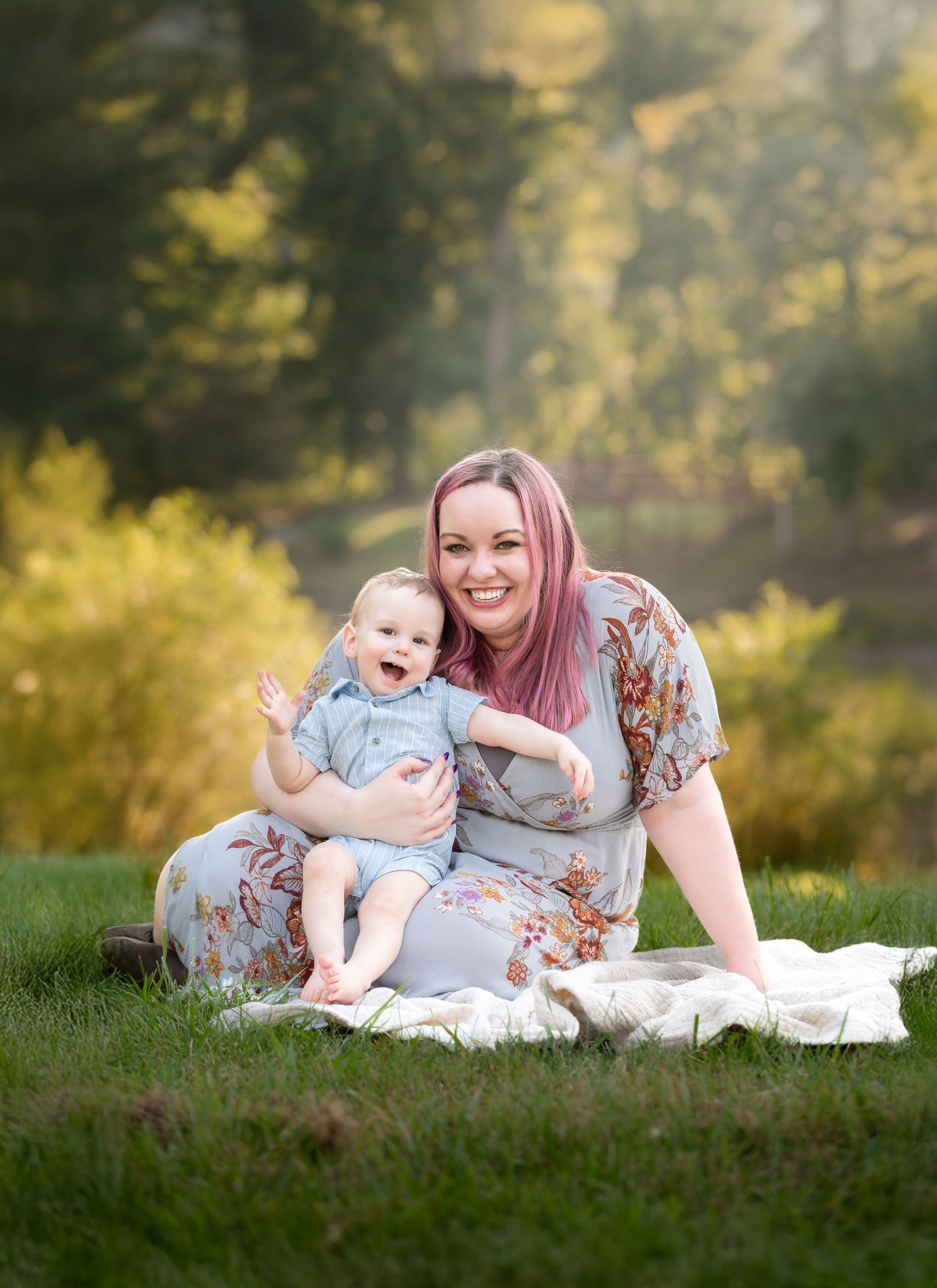 a young mother in a gray floral dress sits on a blanket with her little son, as they both smile for a portrait, with sunlit trees behind