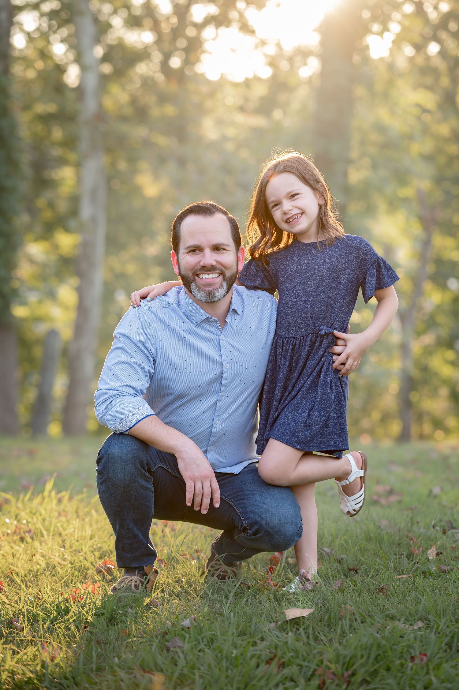 a father crouches beside his young daughter in the grass, with sun shining through trees behind them as they pose for a portrait with smiles