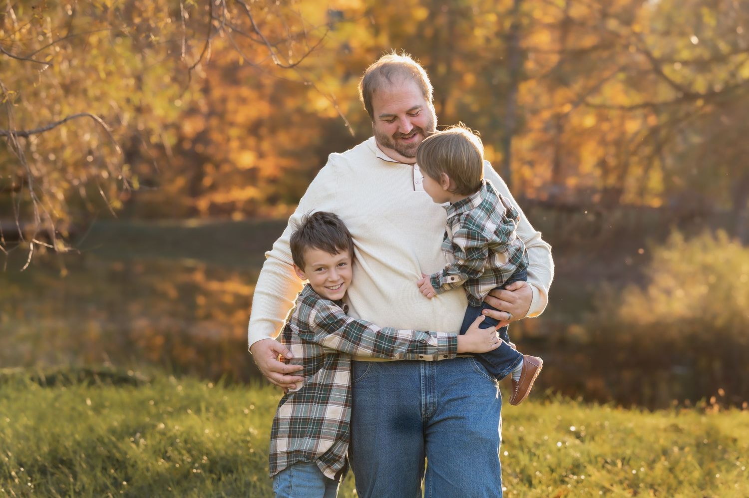 a father hugs and laughs with his two young sons in a field of grass, with autumn leaves in the background, during a portrait event