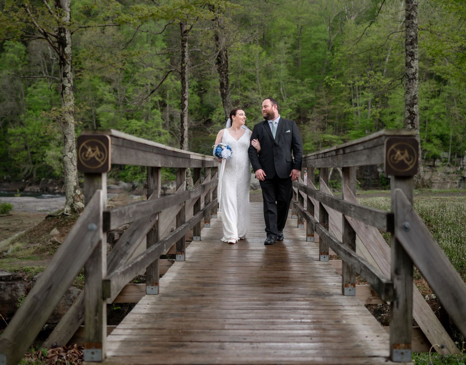 a bride and groom stroll along a wooden bridge hand in hand, with rich green grass and trees all around them, at Valley Falls State Park, WV
