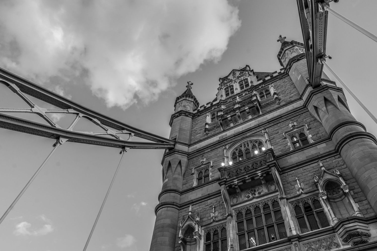 Black and white photo looking up at Tower Bridge London, showing Gothic stonework and suspension cables against cloudy sky.