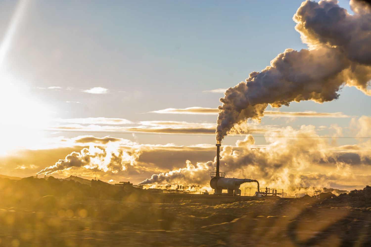 Industrial smokestack emitting large plumes of smoke against a golden sunset sky with dramatic clouds.