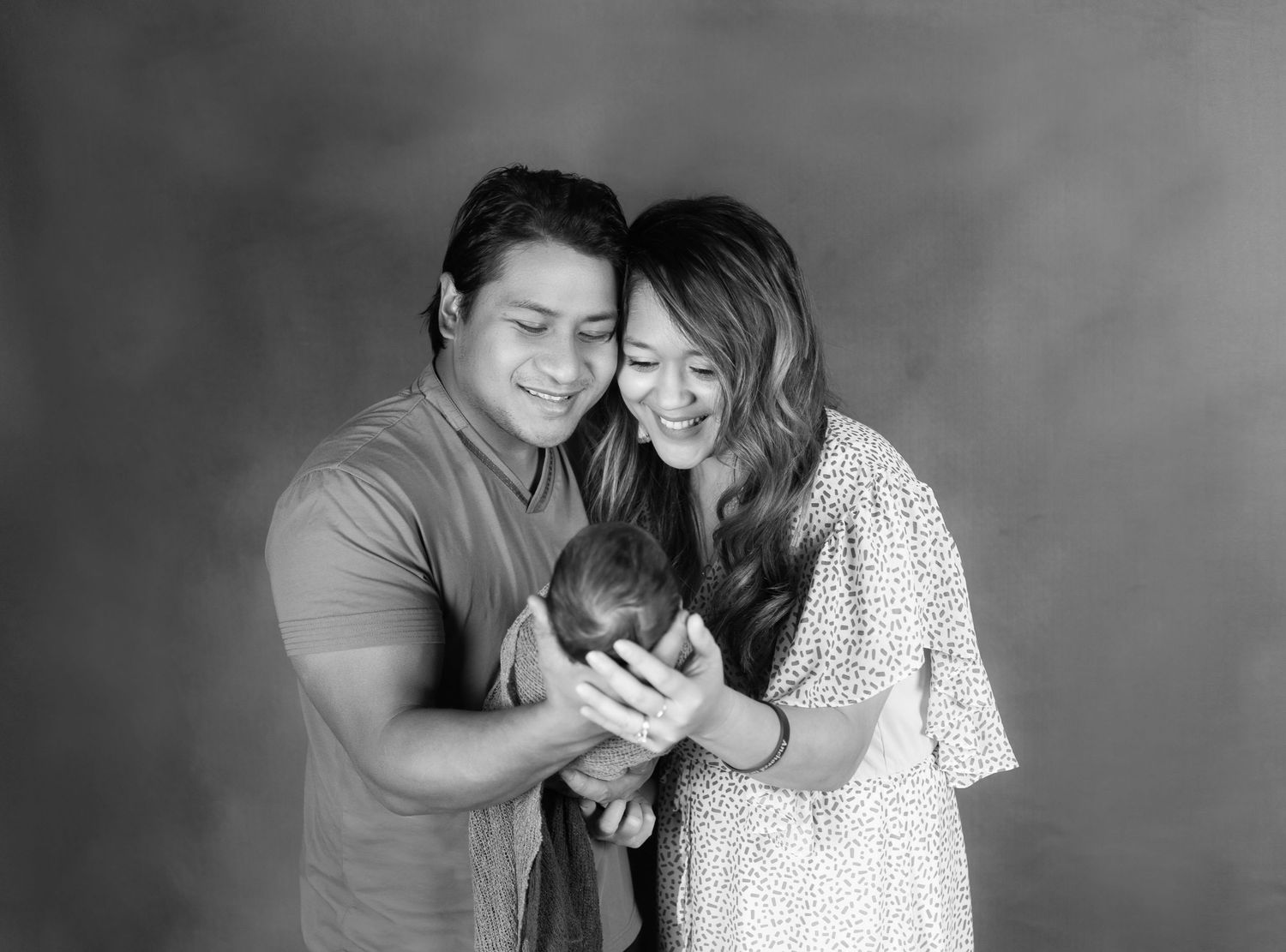 Black and white studio portrait of smiling parents lovingly holding their newborn baby together.
