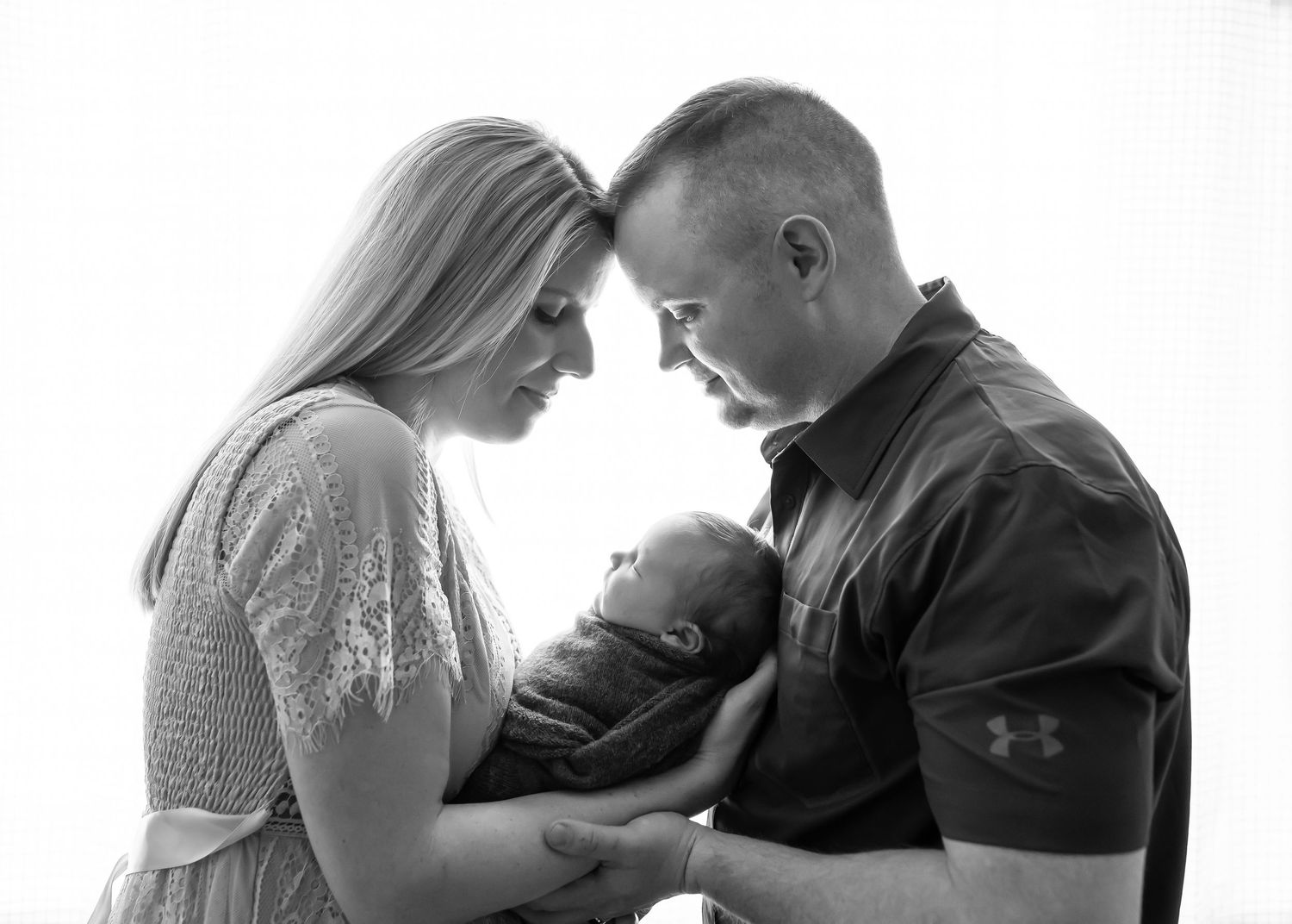 Black and white photo of parents tenderly holding their newborn baby together in a bright studio setting.