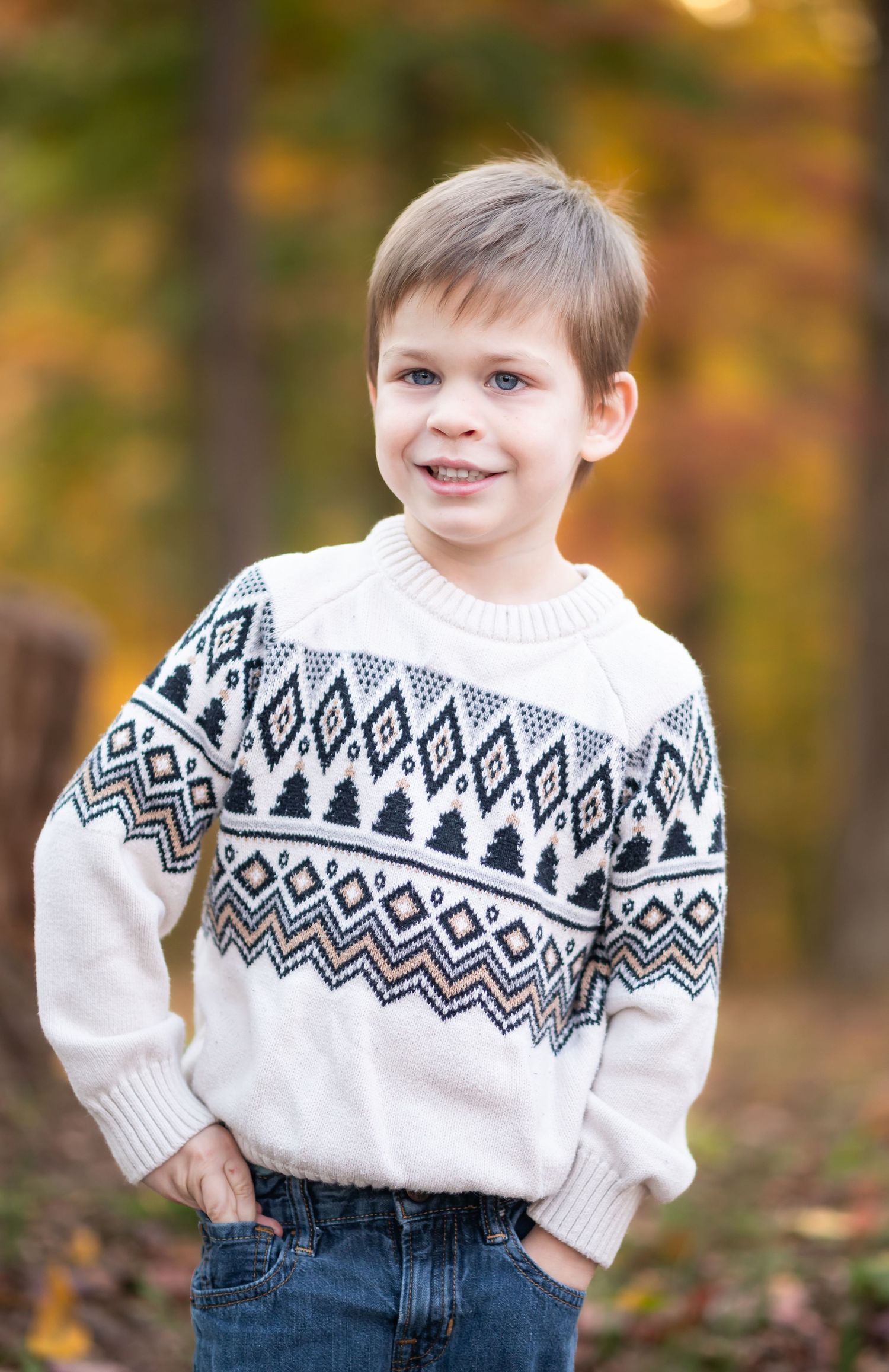 a little boy in a white argyle sweater poses and smiles with hands in pockets and fall foliage behind him in the background