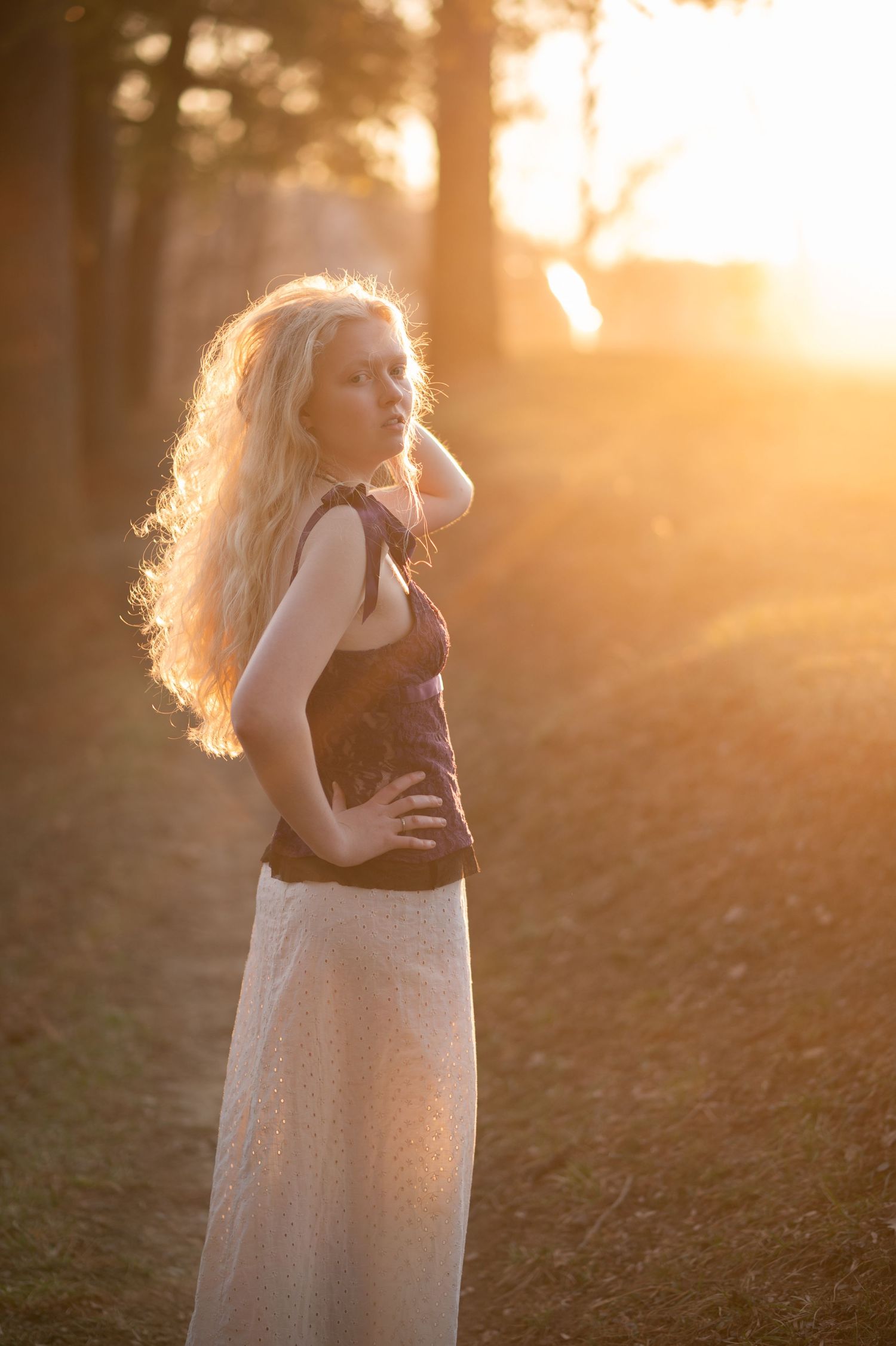 sun-drenched golden hour portrait of a teen girl with long blonde hair, posing with her hand on her hip while the sun shines behind her
