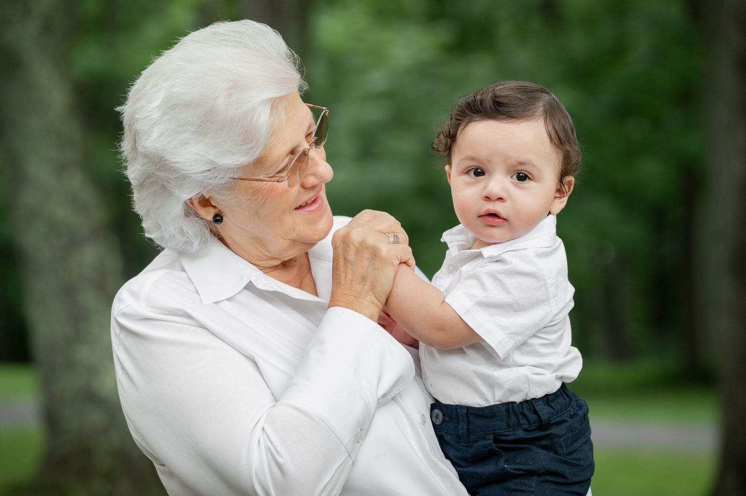 a grandmother in a white shirt with white hair holds her little grandson, holds his hand and smiles at him as he looks at the camera