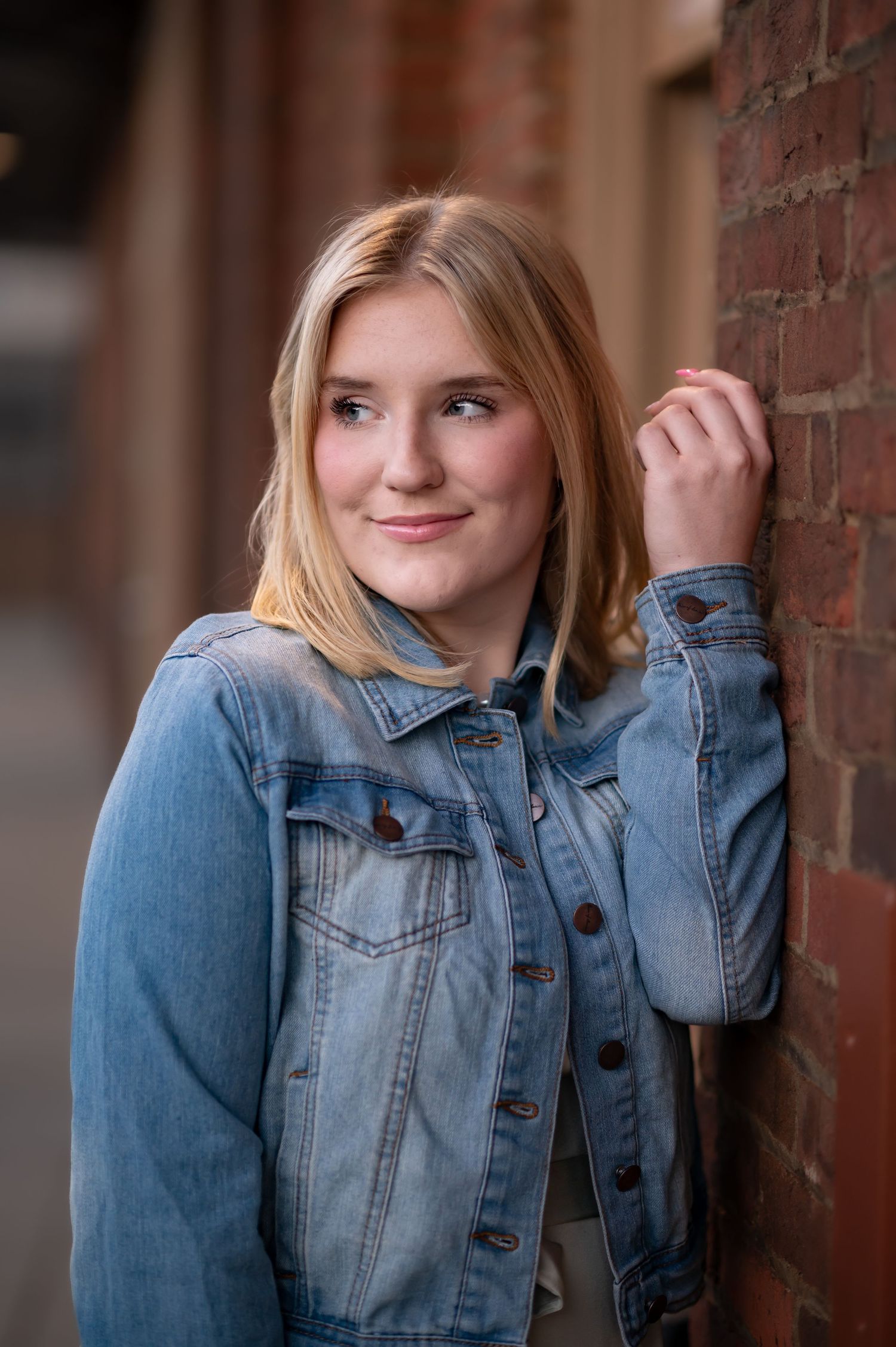 closeup portrait of a blonde high school student in a jean jacked, leaning against a brick wall and looking away from the camera