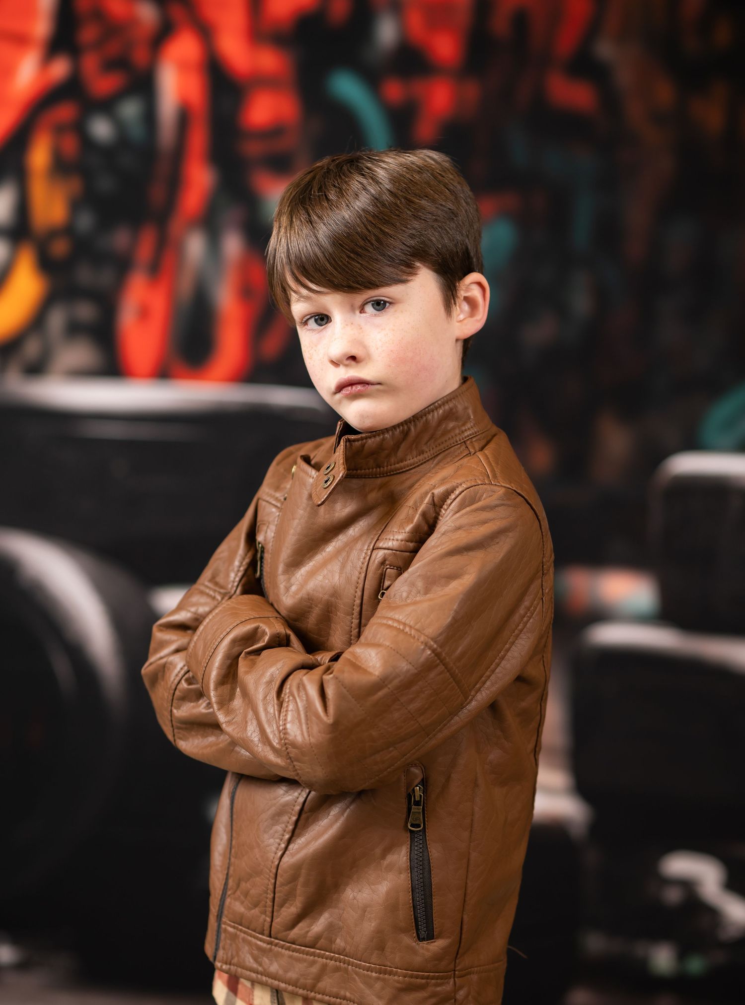 a brown haired boy in a brown leather jacket poses with arms crossed and a solemn expression in an alleyway stacked with old tires