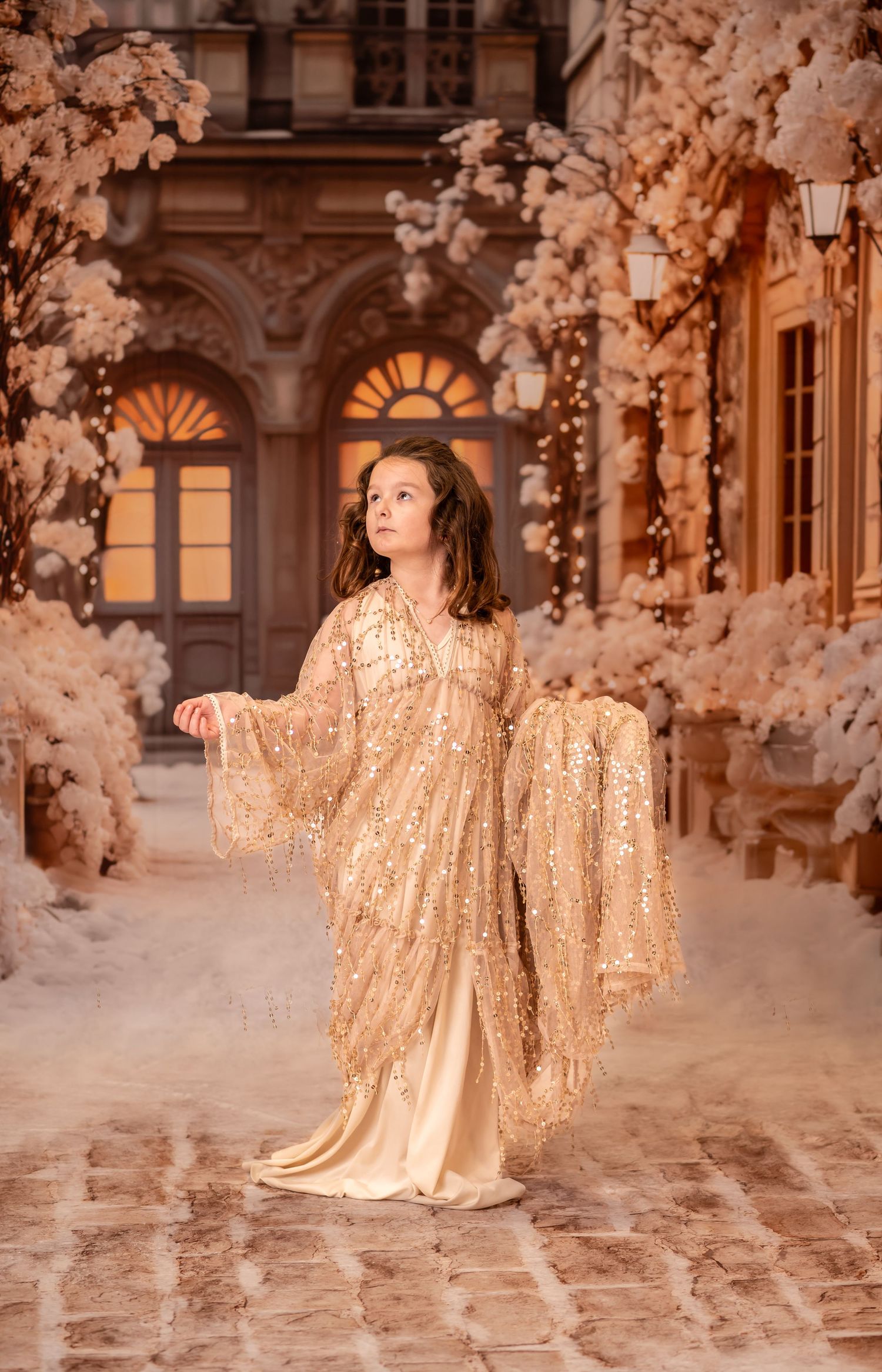 a little girl walks down a snowy brick street in a dramatic gold sequin dress, with soft beige and ivory Christmas decor in the background