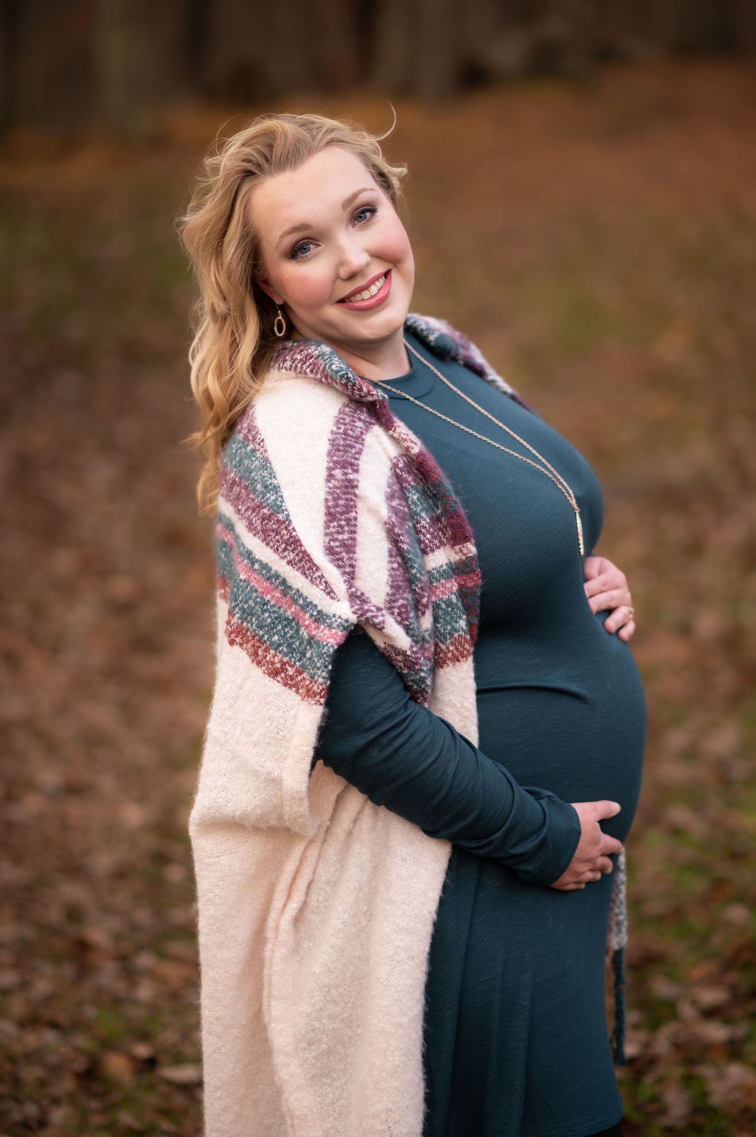a pregnant woman in a long teal dress and a cream sweater poses with her hands on her belly and smiles for a picture