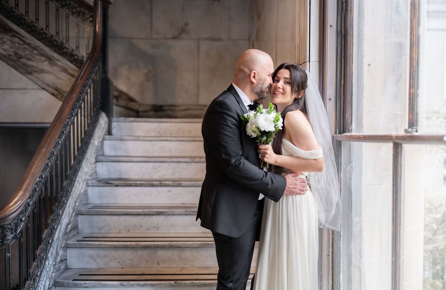 a bride and groom in traditional wedding attire pose for a portrait by a large old window on a marble staircase