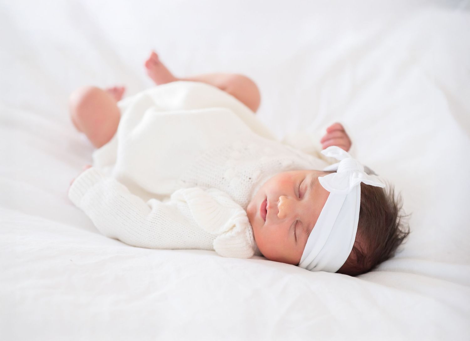 Newborn baby girl sleeping peacefully in white outfit and bow headband on soft white bedding.