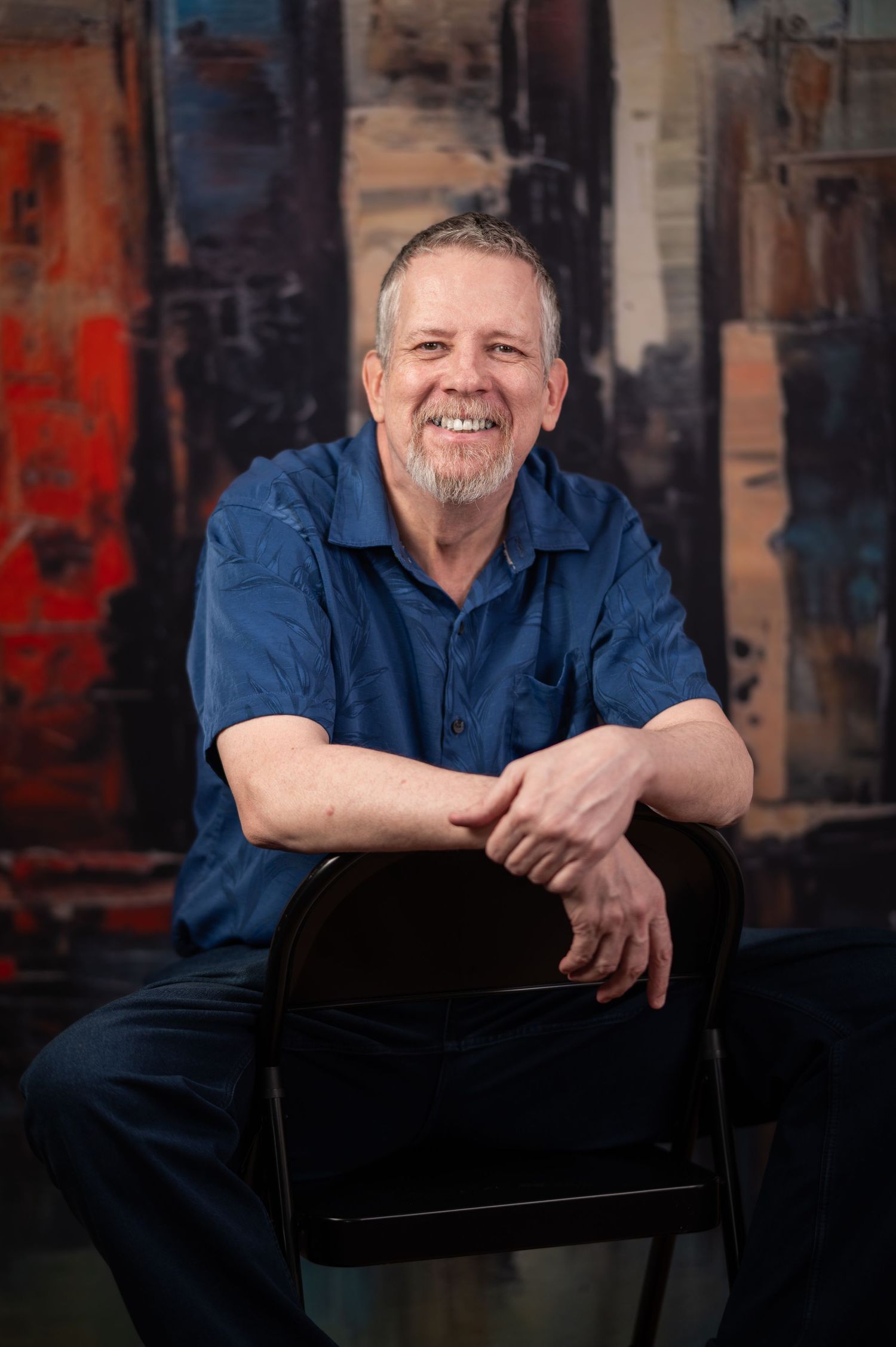 A bearded man leans his arms on the back of a chair and smiles for a portrait, with a bright abstract painting behind him.