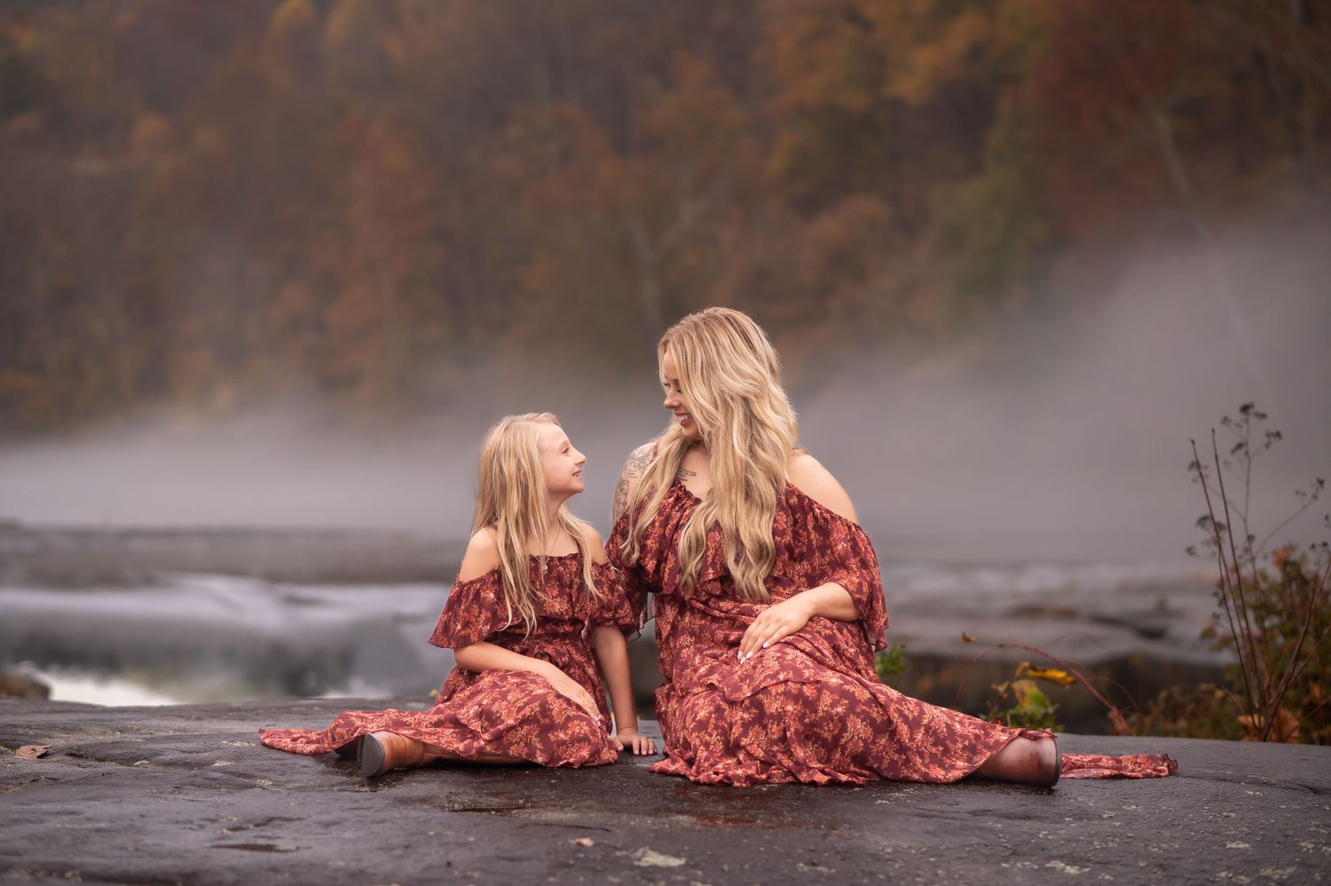 Outdoor autumn portrait of a blonde mother and daughter in matching red floral dresses, posing seated on a large rock at Valley Falls, WV
