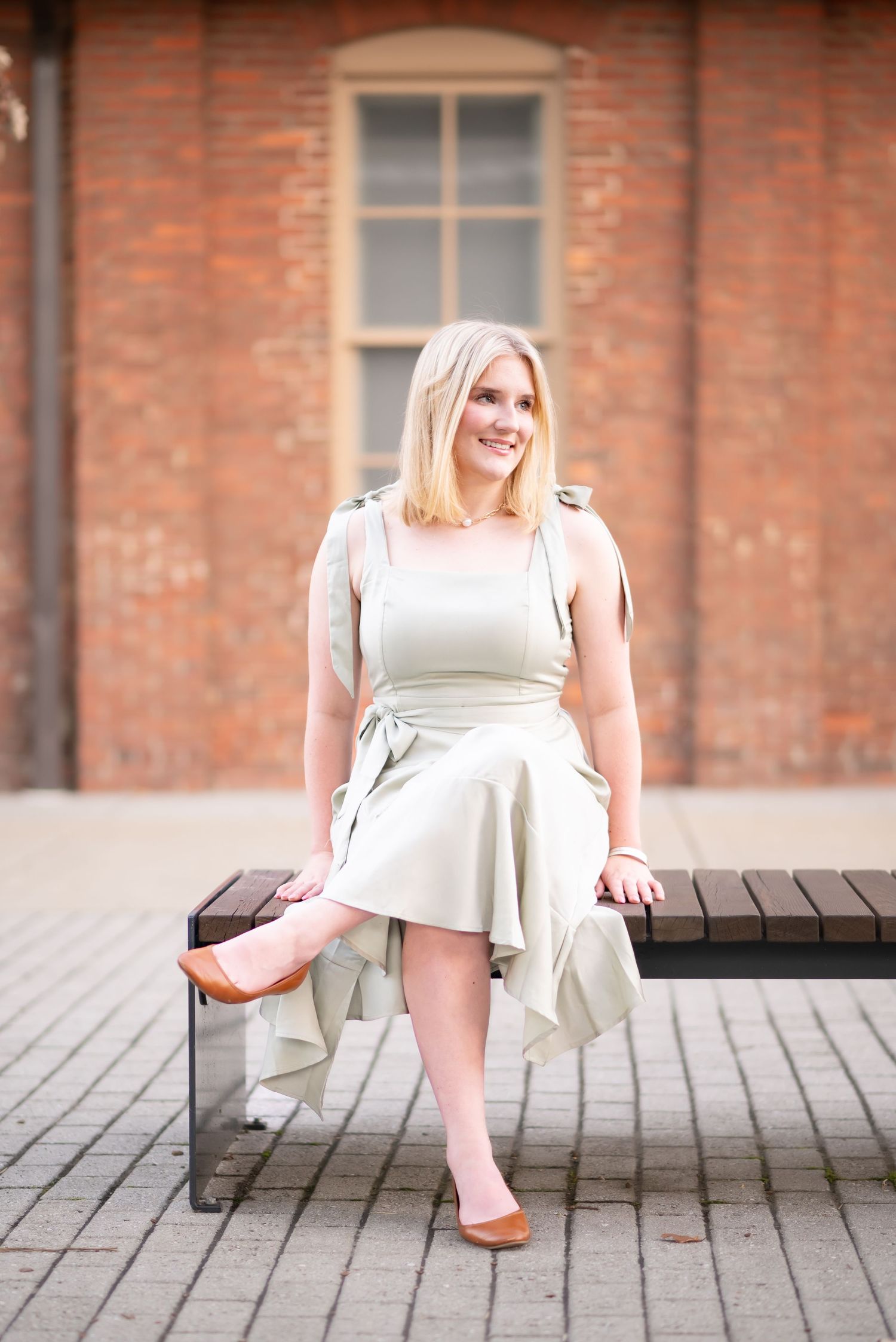 a young woman in a green dress poses for a picture on a slatted bench , with an old brick bus depot in the background