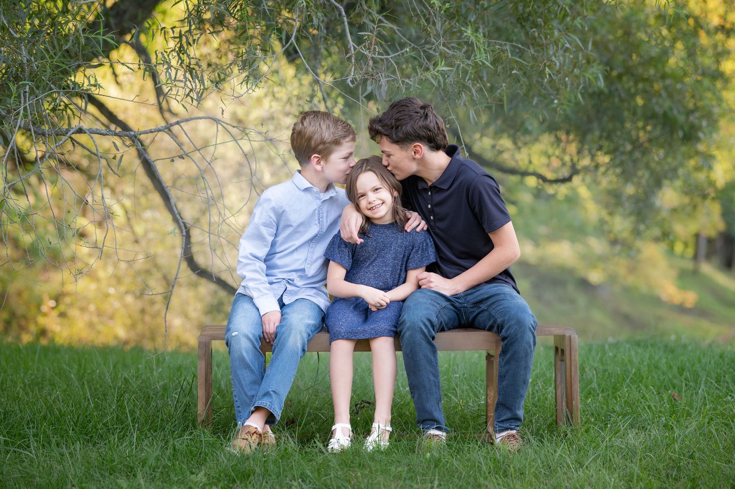 two brothers sit on a bench in the grass with their little sister in the middle, and playfully kiss her on the head as she laughs