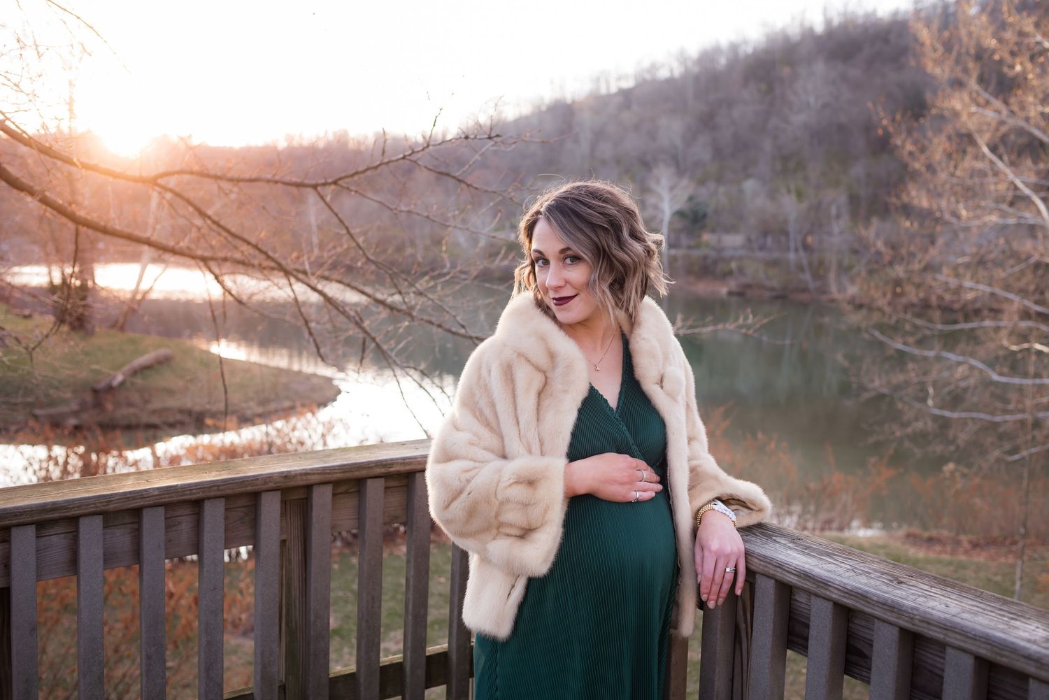 a lovely pregnant woman in a green dress and beige fur cape poses against a wooden railing with a river and wintery mountains behind her