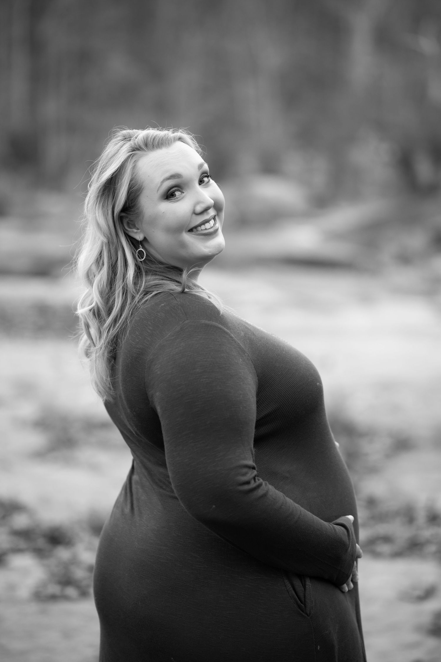 black and white portrait of a pretty blonde pregnant lady in a black sweater dress smiling back over her shoulder at the camera