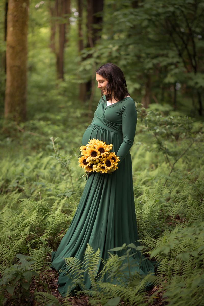 a pregnant woman in a long dramatic green dress poses with a bouquet of sunflowers in a green patch of ferns in the woods