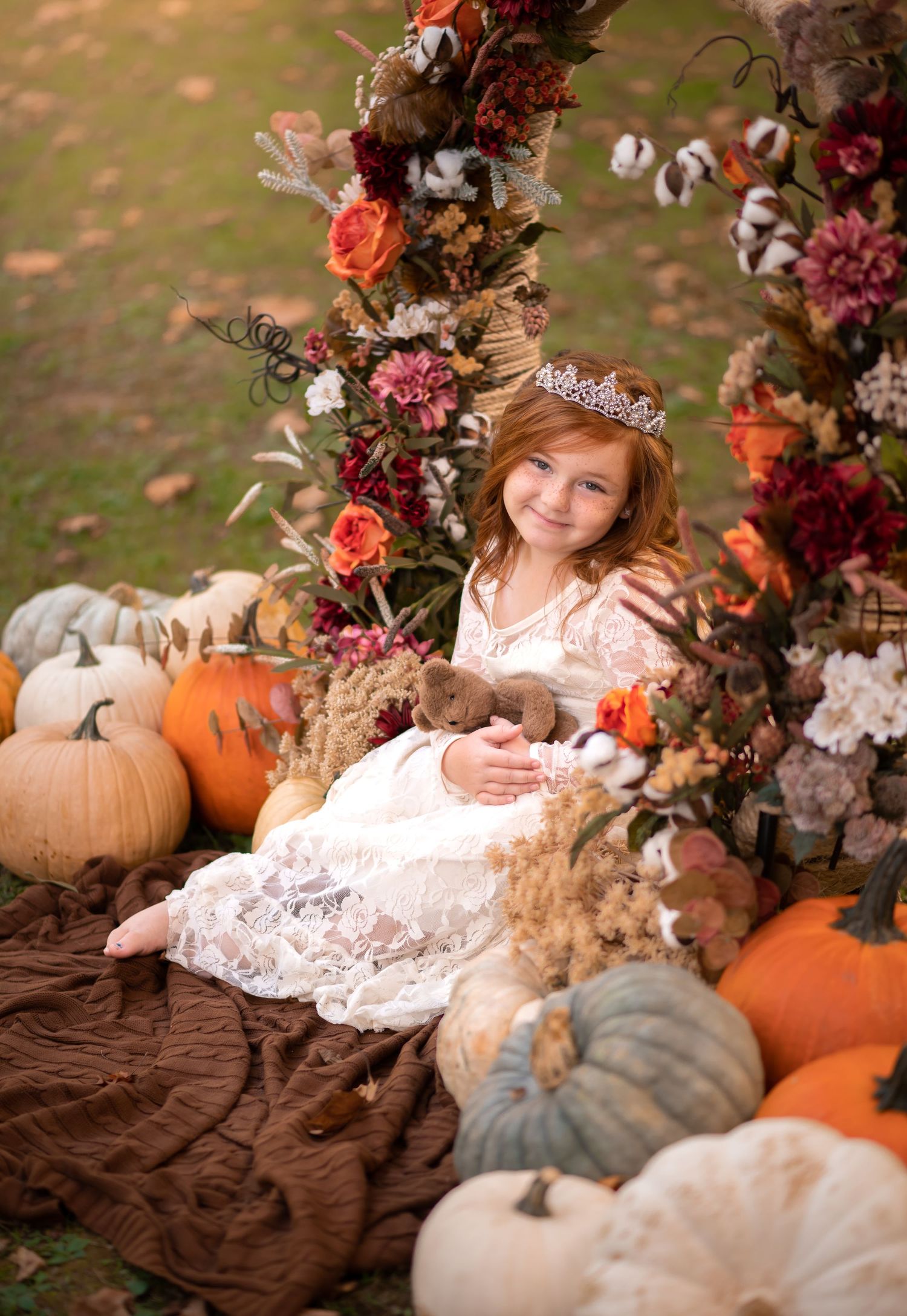 outdoor autumn portrait of a little red haired girl in a white dress, sitting in an autumn floral arch with pumpkins all around