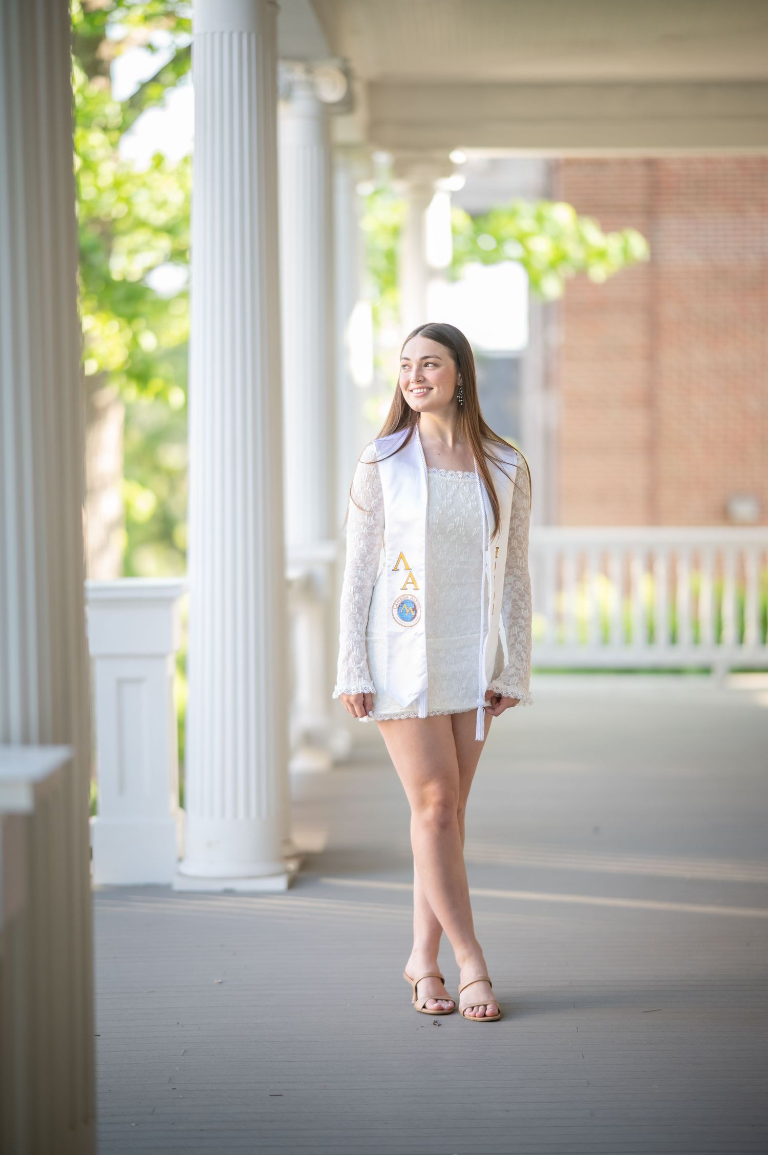 a WVU graduate student in a white dress poses for a senior portrait on a gray porch with white pillars in the background
