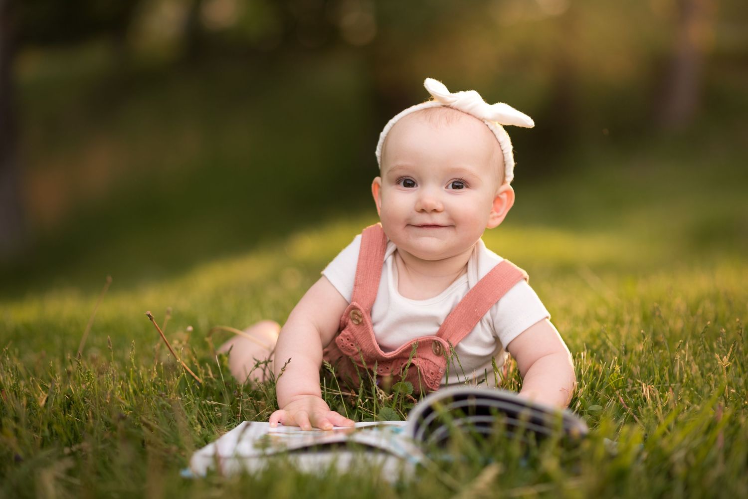 a cute toddler girl in orange overalls lies in the grass and looks at a book