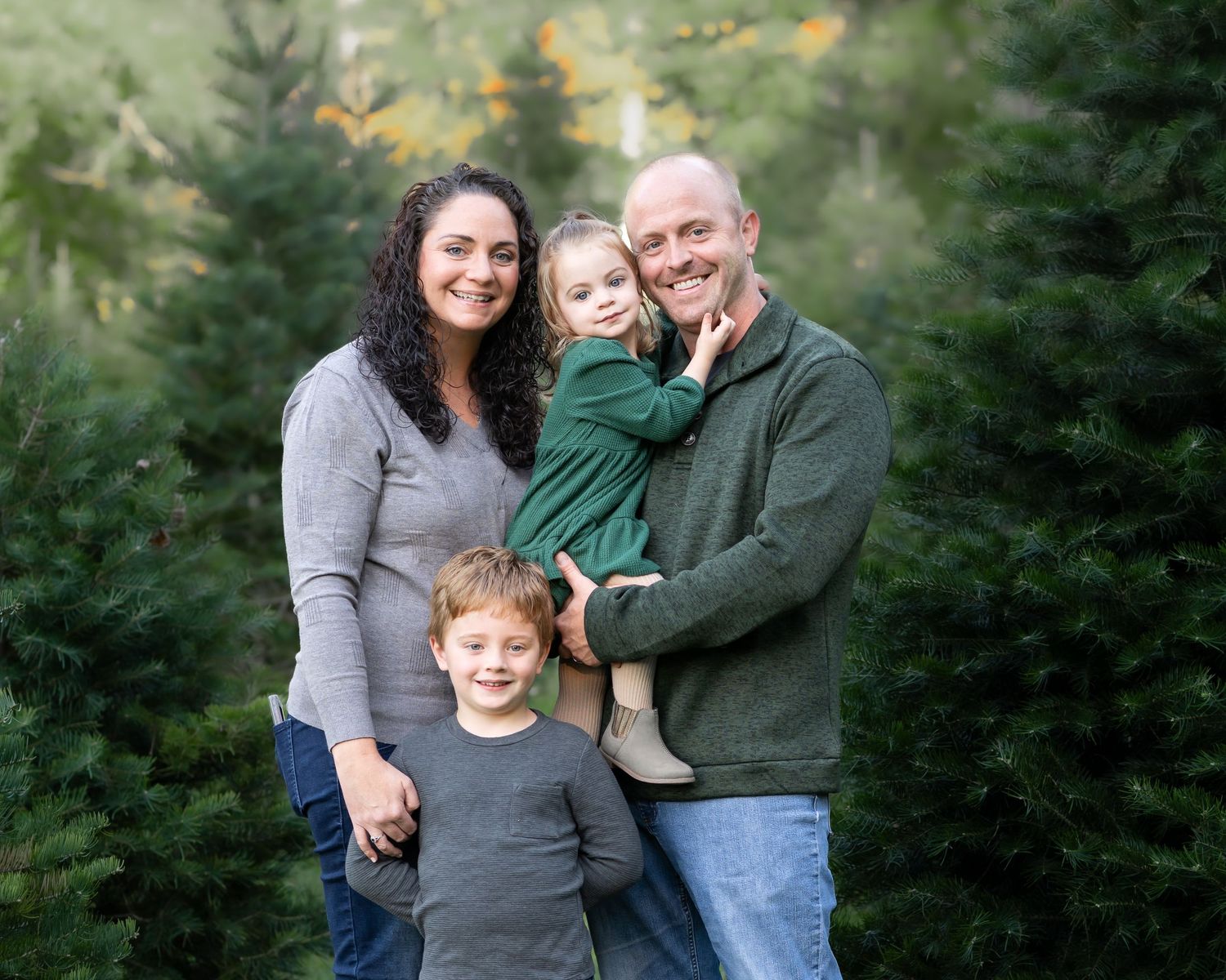 a family of four poses and smiles for a Christmas portrait in a lush green tree farm in Fairmont, WV, surrounded by Christmas trees.
