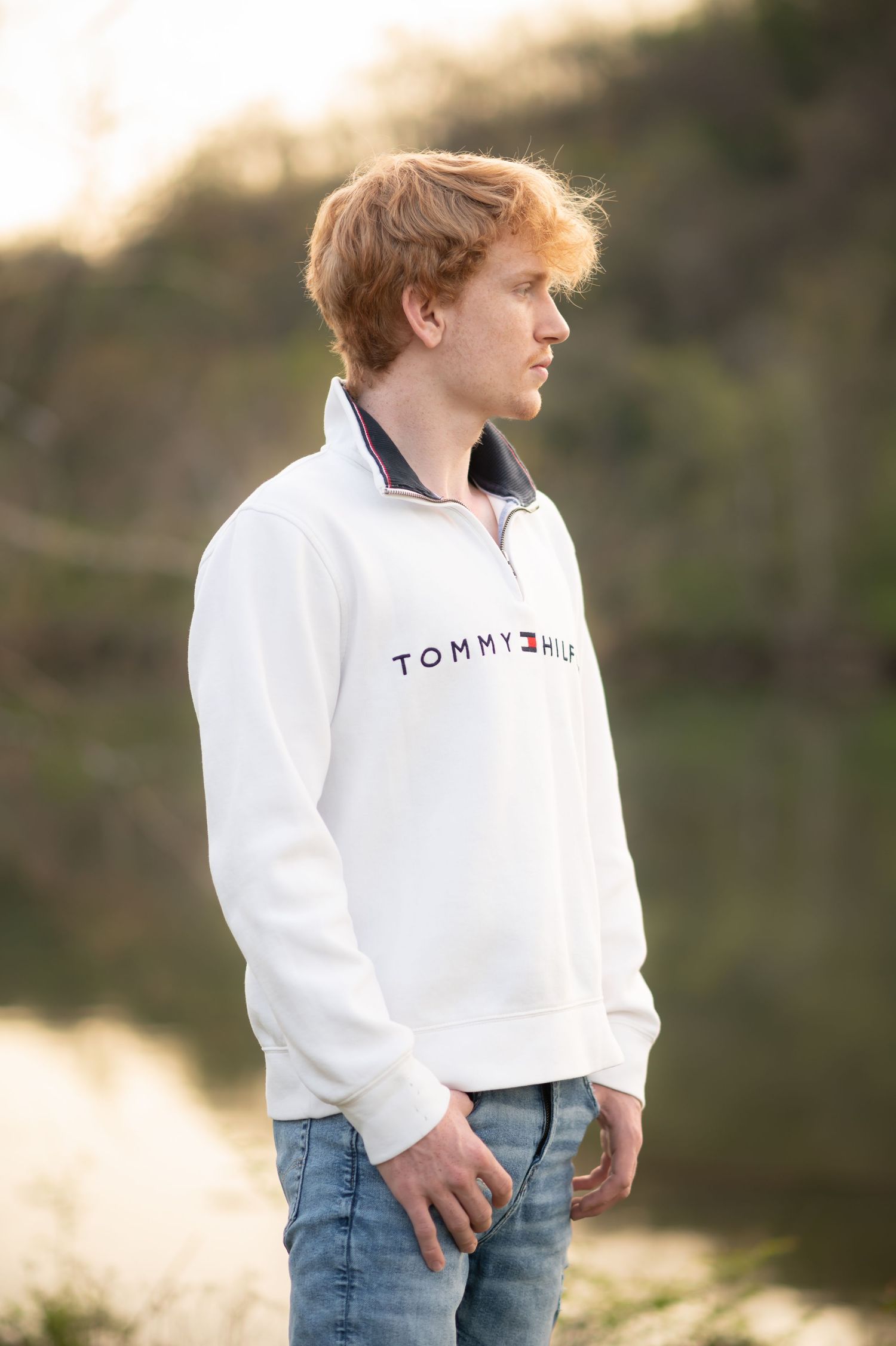 senior portrait of a young man in a white sweater, looking out over a river as the sun sets behind him