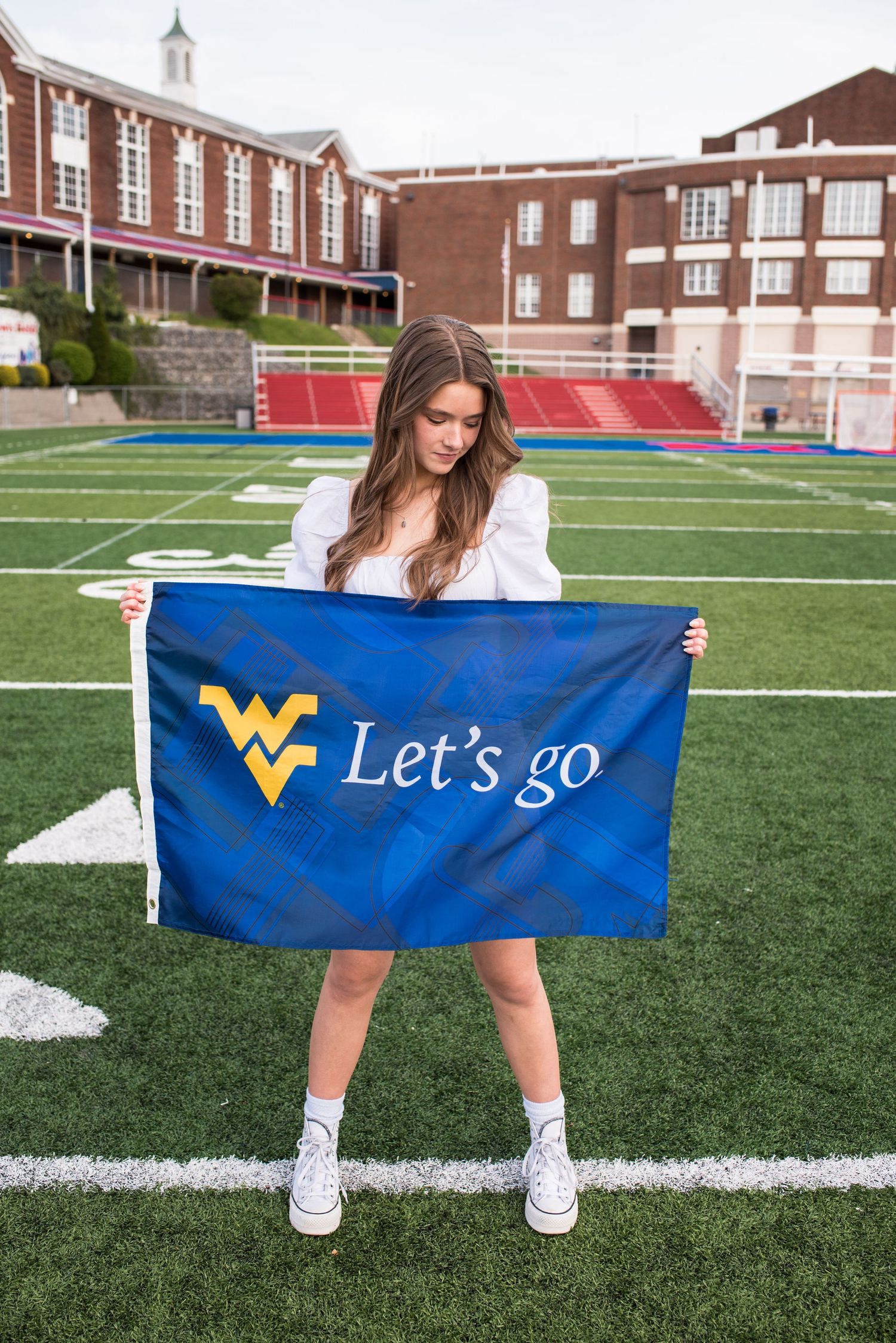 a high school student in a white dress and sneakers holds a blue and gold WVU flag while standing on the MHS football field