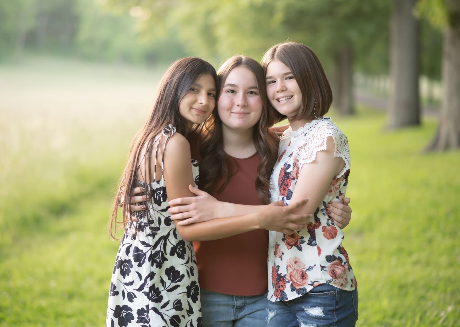 three young women sisters pose close together and smile for an outdoor portrait in a sunlit field with trees in the background