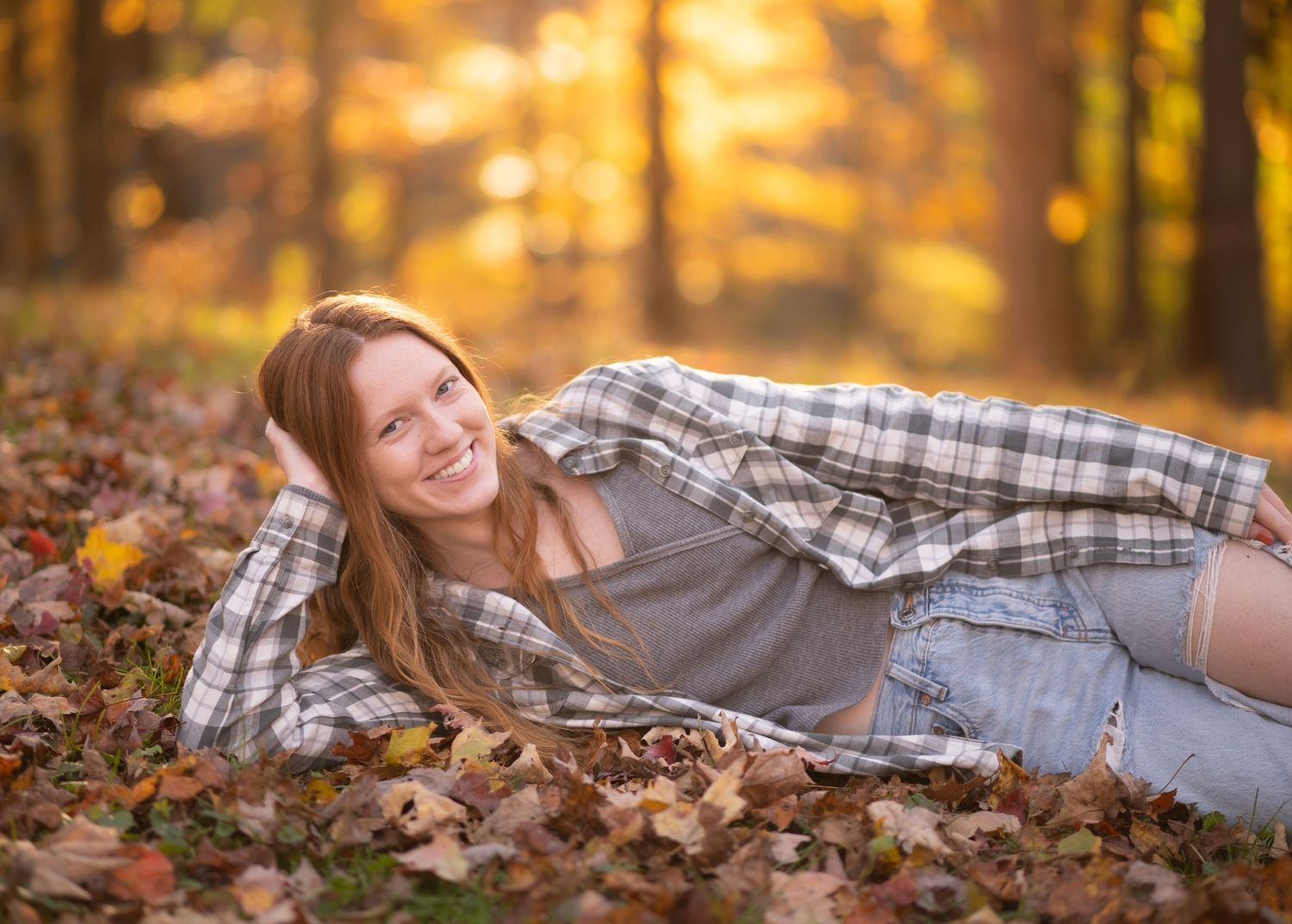 a happy high school student with long red hair and a plaid shirt lounges on the ground, surrounded by brightly colored autumn leaves