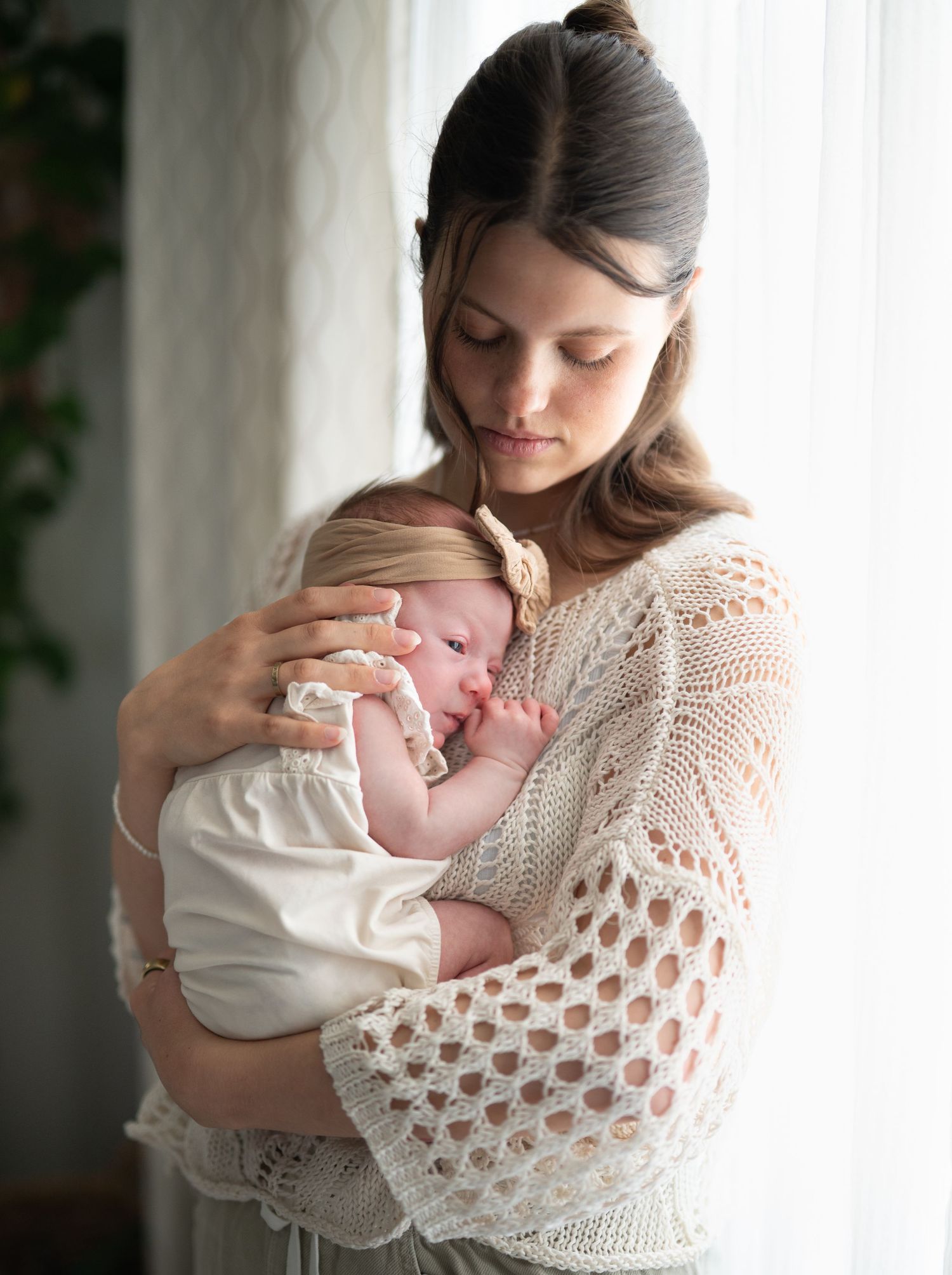A young mother in a cream crochet sweater tenderly holds her sleeping newborn baby near a bright window.