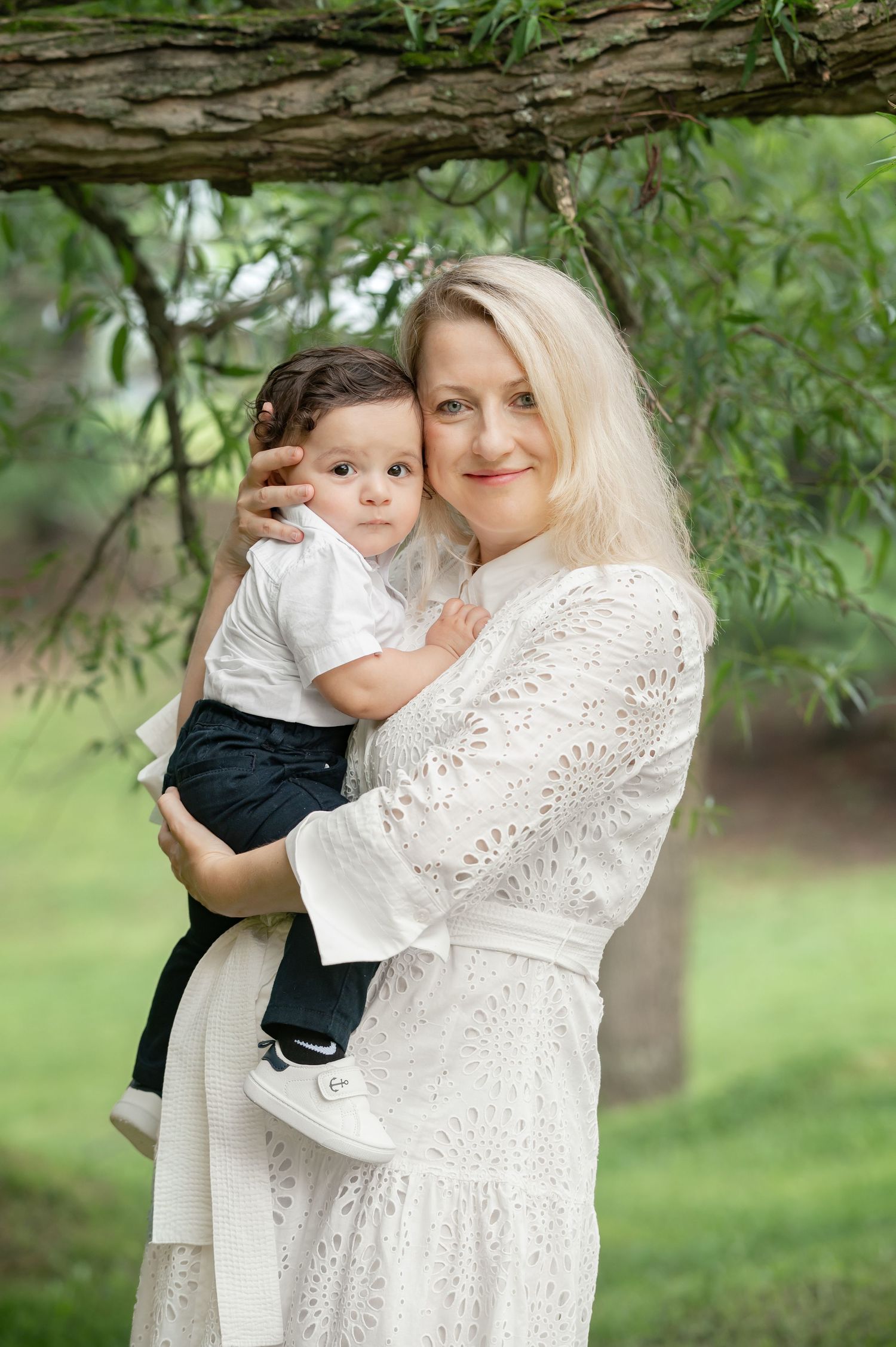 a mother in a white dress holds her little son, with faces close together, and trees in the background