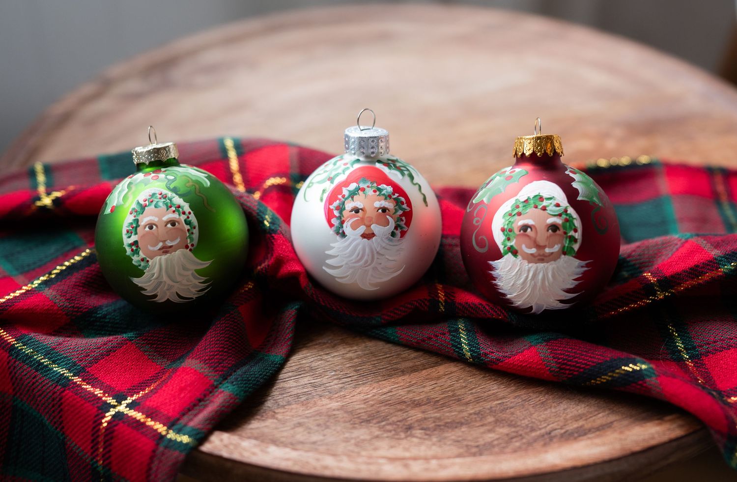 Product image of three Santa Claus Christmas ornaments on plaid ribbon and wooden board, featuring green and white holiday ball decorations.