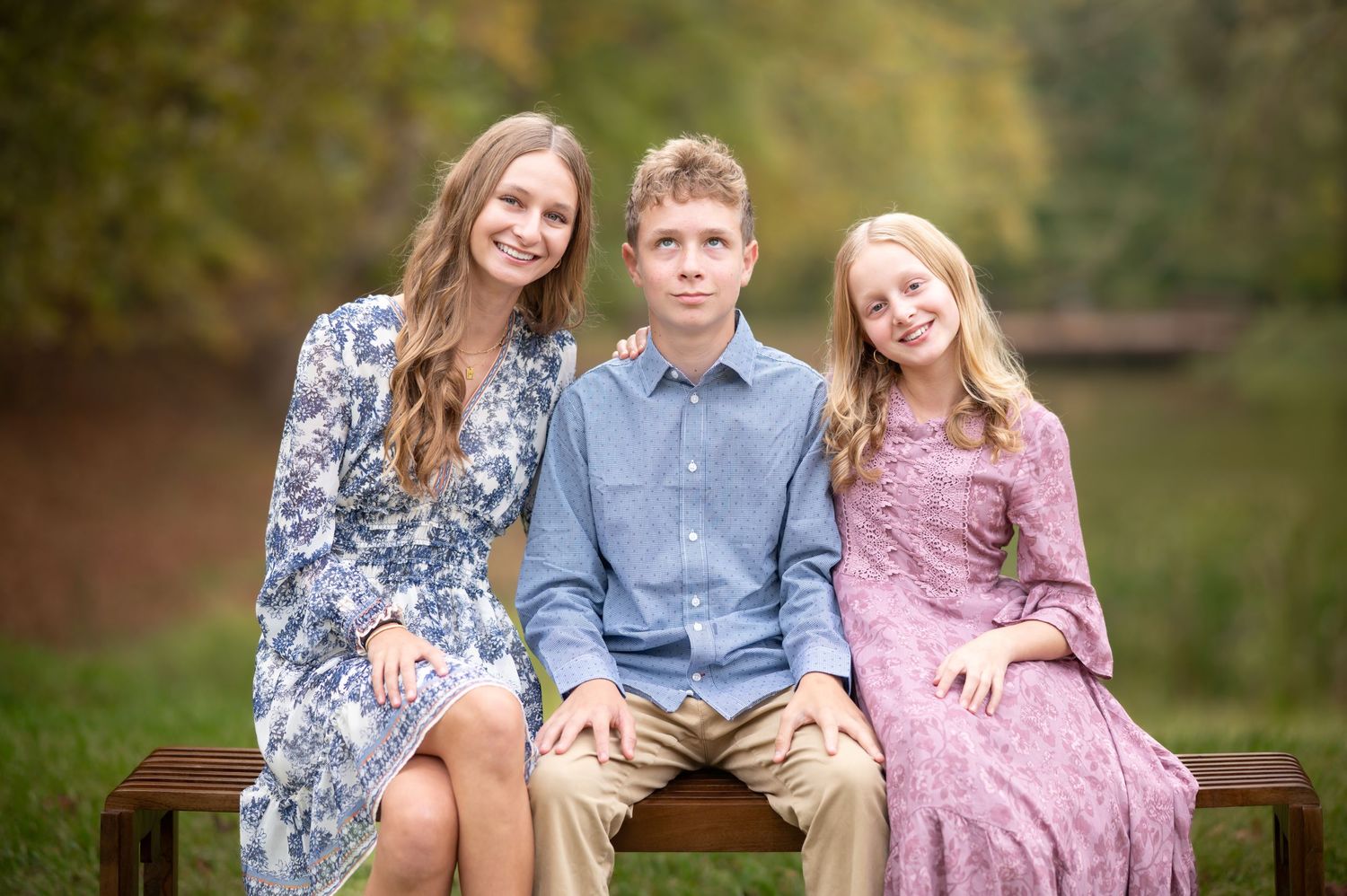 two sisters sit on a bench with their brother sitting between them. They smile for the camera while he rolls his eyes