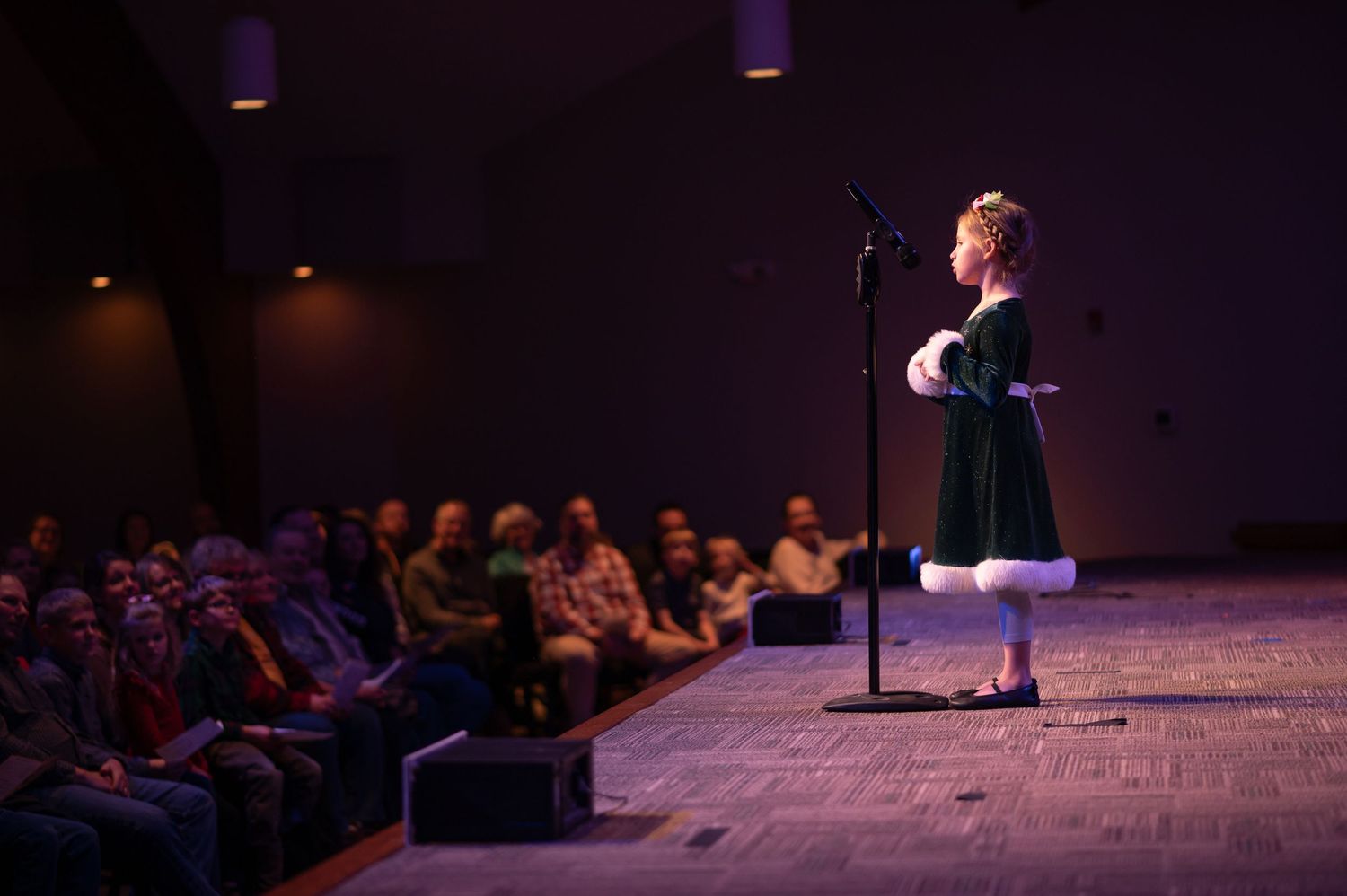 a little girl in a green velvet dress with white fur cuffs stands nervously on a lit stage in front of a microphone in front of an audience