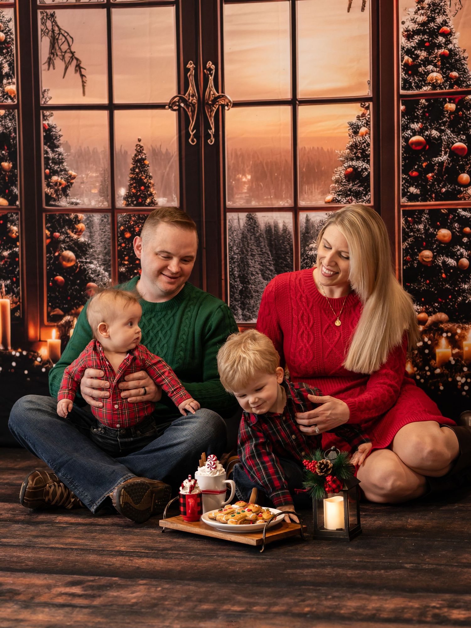 a family in green and red Christmas sweaters sits on a wood floor with a tray of hot cocoa and a beautiful sunset Christmas scene behind