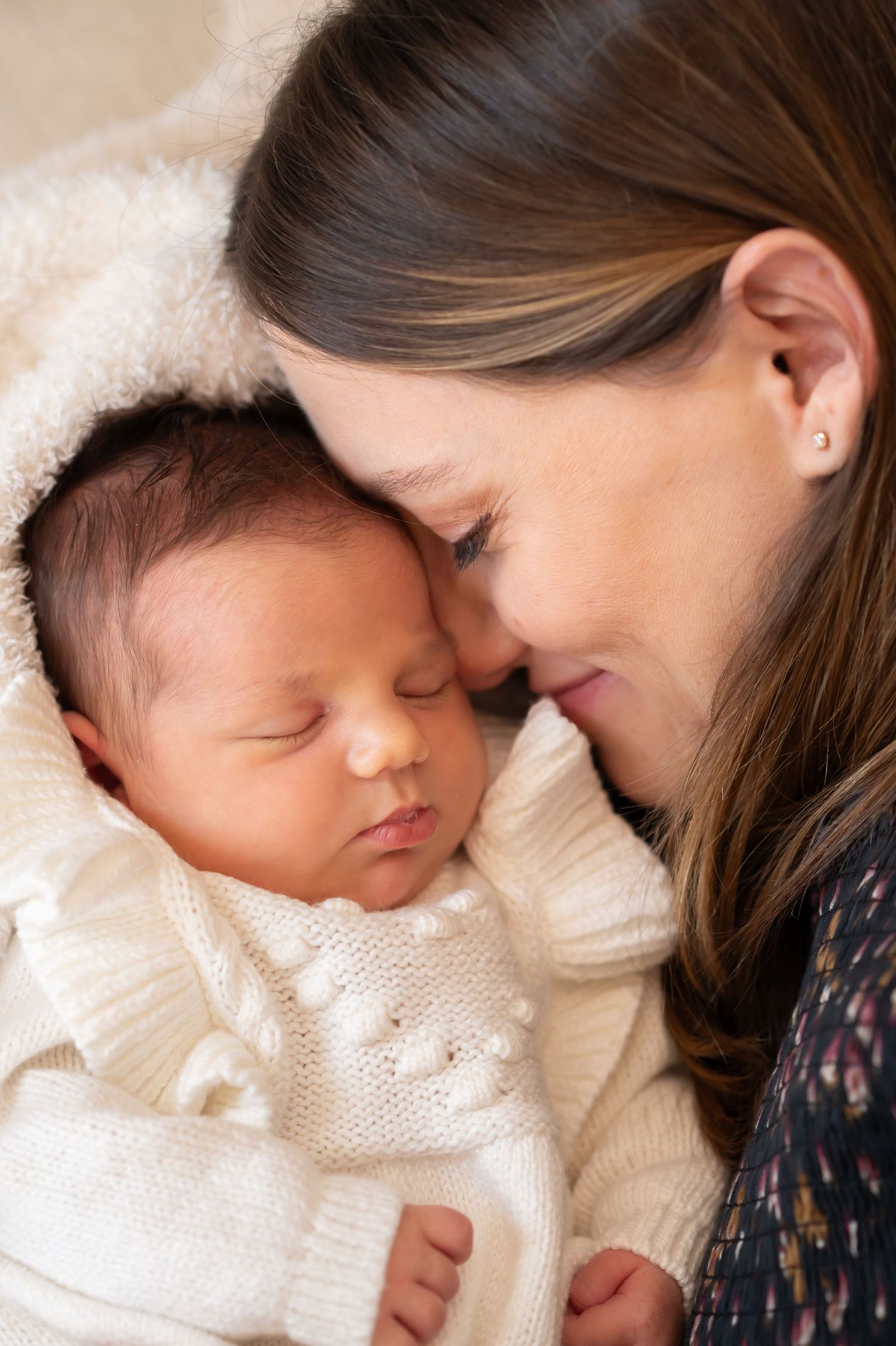 A tender moment as a young mother gently touches foreheads with her sleeping newborn baby dressed in white knit outfit.